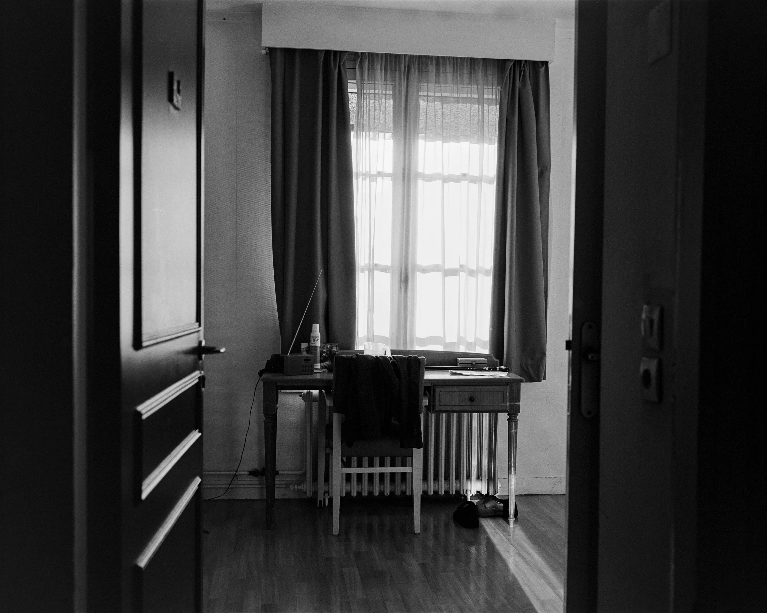 Une vue d'un bureau dans une pièce, vue à travers une porte entrouverte. Le bureau est contre une fenêtre avec des rideaux. Il y a une chaise avec un vêtement dessus, et divers objets sur le bureau, comme une bouteille, des documents et une radio.