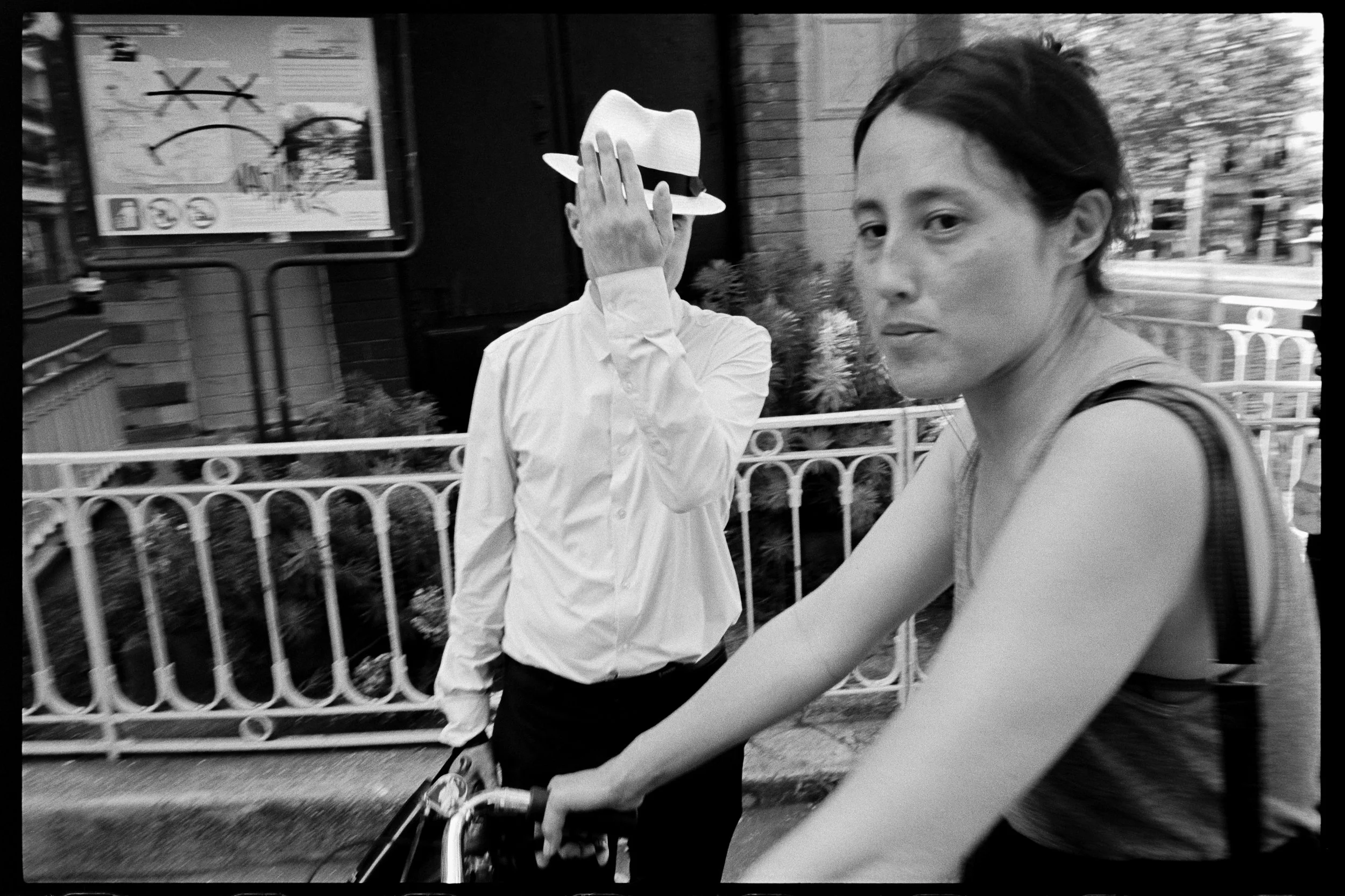 Une femme à vélo devant un mur avec une affiche, un homme en chemise blanche avec un chapeau blanc et une clôture en fer forgé en arrière-plan.