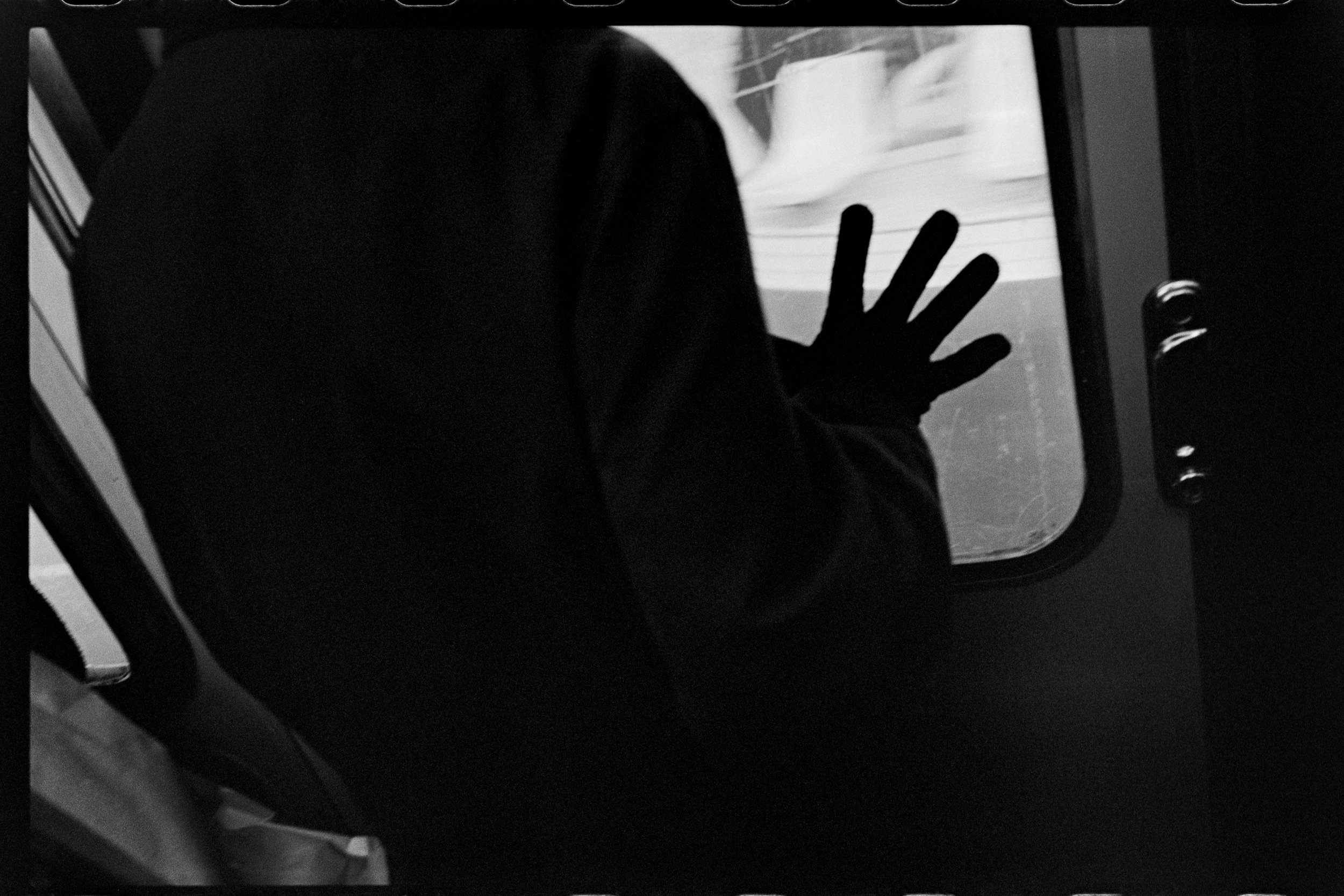 Une personne en vêtements sombres regarde par une fenêtre d'un véhicule, la main posée sur la vitre.