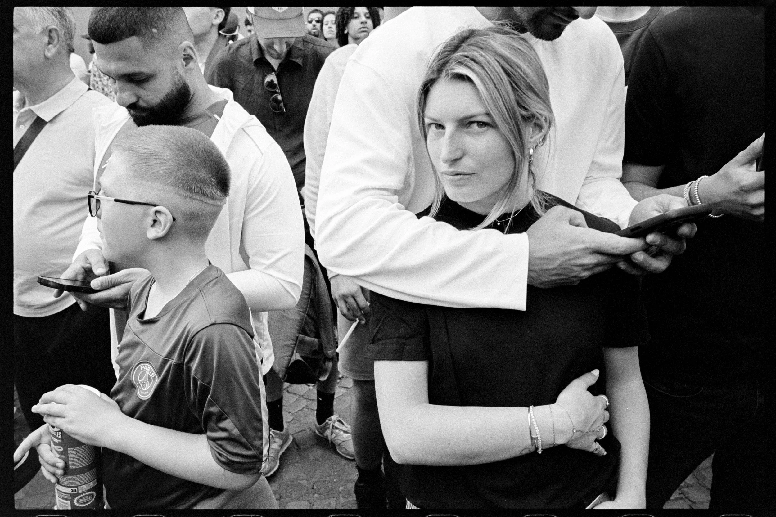 Groupe de personnes dans une foule, certaines utilisant leur téléphone portable, une femme regarde l'appareil avec expression concentrée, en noir et blanc.