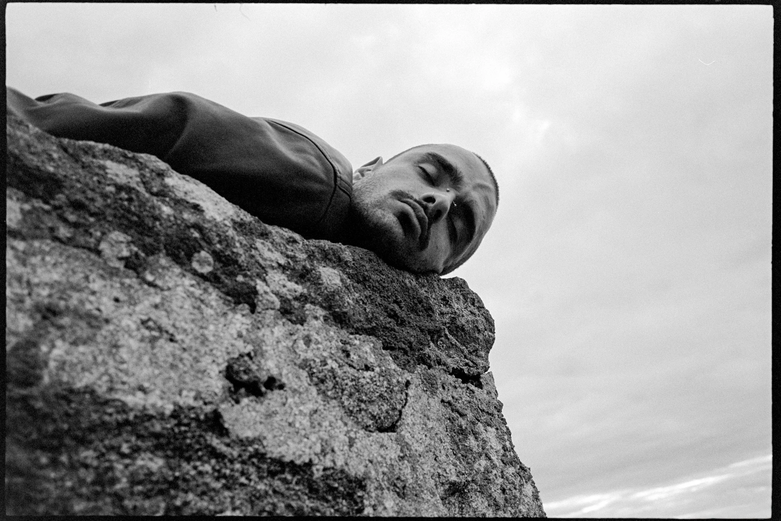 Un homme avec un visage pâle et une barbe regarde vers le bas, allongé sur une roche.
