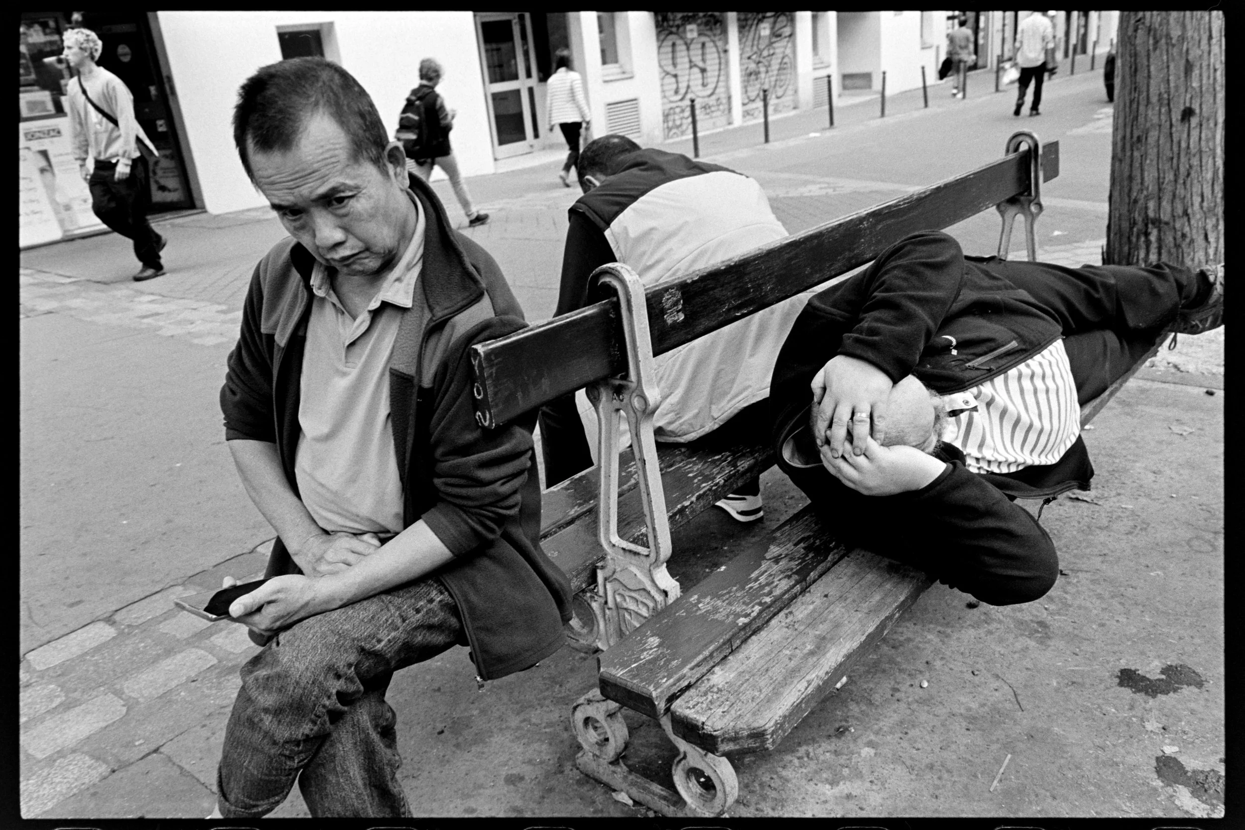 Trois personnes sur un banc dans une rue urbaine, deux assises et une allongée. L'une des personnes assises regarde son téléphone, une autre se tient avec la tête baissée et la troisième repose la tête dans ses mains.