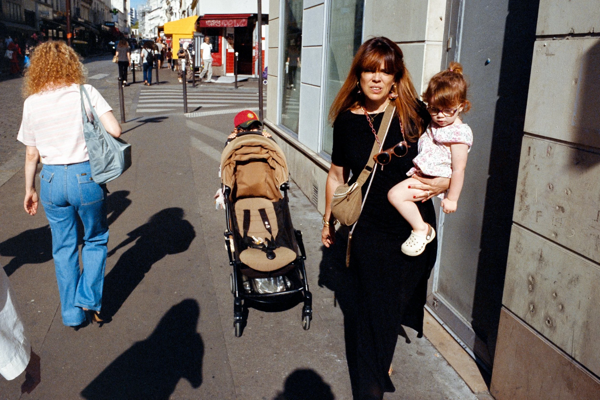 Une femme portant une robe noire tient une petite fille dans ses bras dans une rue animée. Il y a une poussette à côté d'elles, d'autres personnes marchent, et des bâtiments urbains en arrière-plan.
