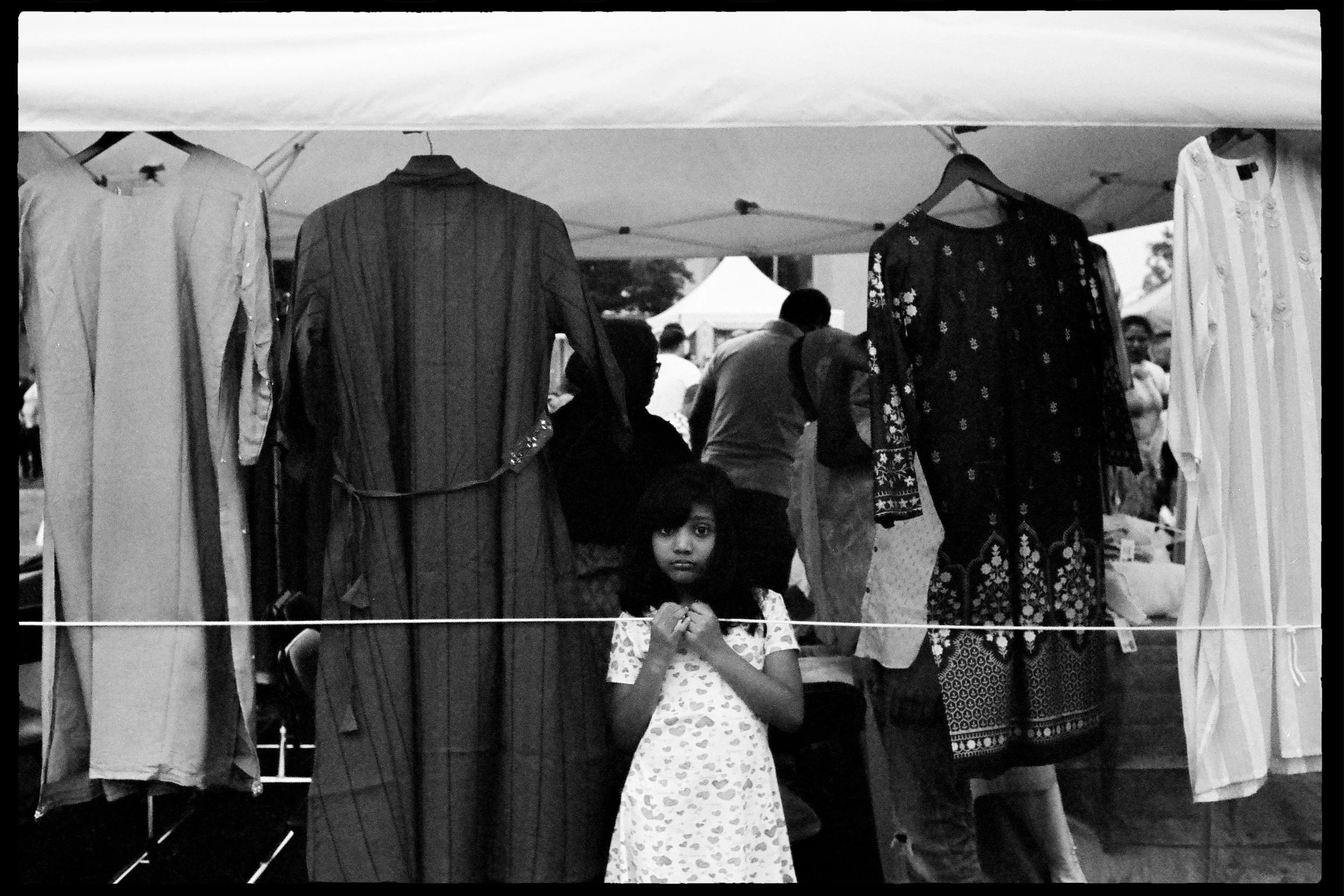 Un stand avec des vêtements suspendus, y compris deux robes longues, une noire à motifs et une blanche rayée. En face du stand, une jeune fille regarde l'objectif, tenant ses mains près du col de sa robe. Des personnes sont visibles en arrière-plan, 
