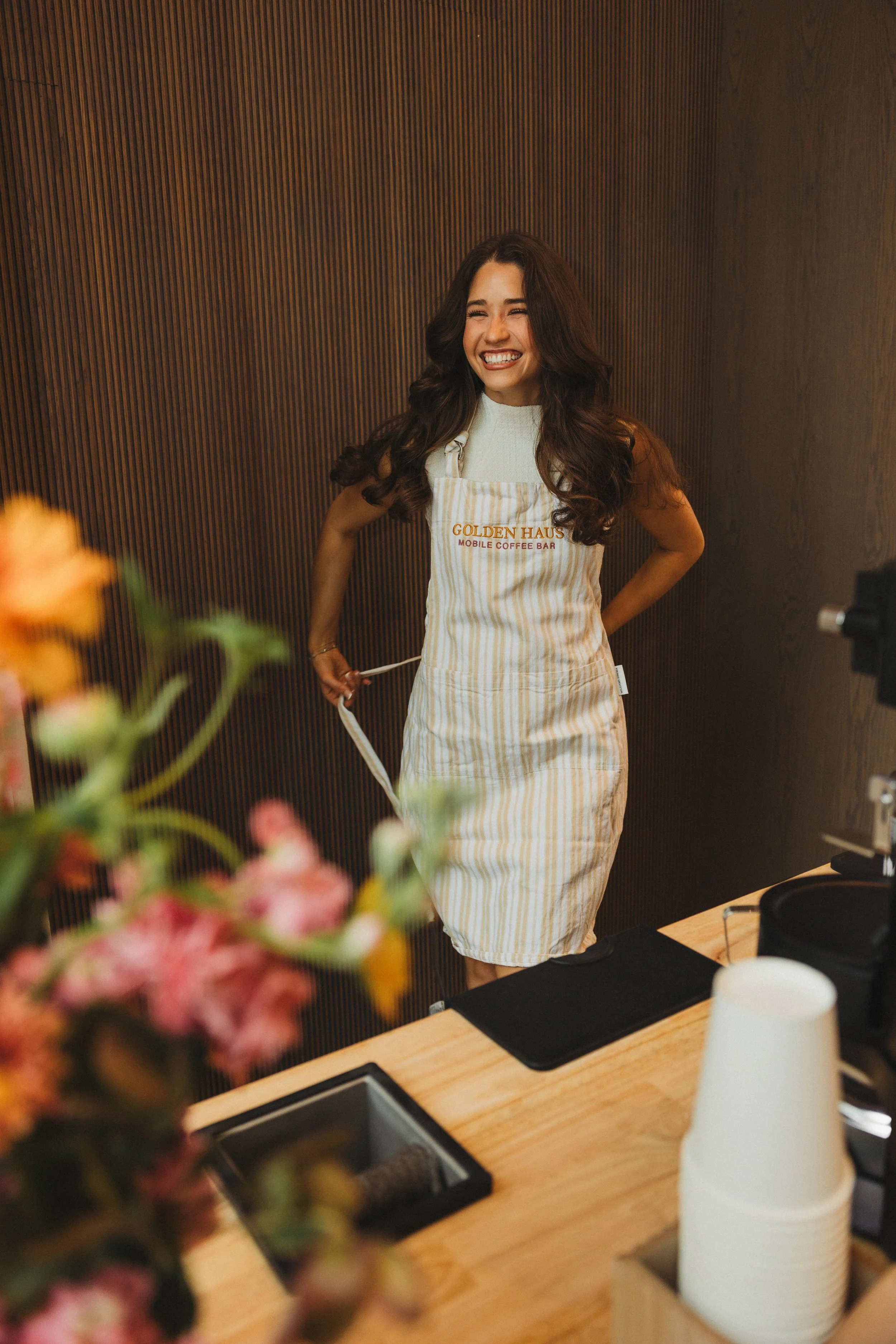 A smiling woman wearing a white apron with yellow and beige vertical stripes, standing behind a coffee bar with a wooden counter, flowers in the foreground, and a dark wooden wall in the background.
