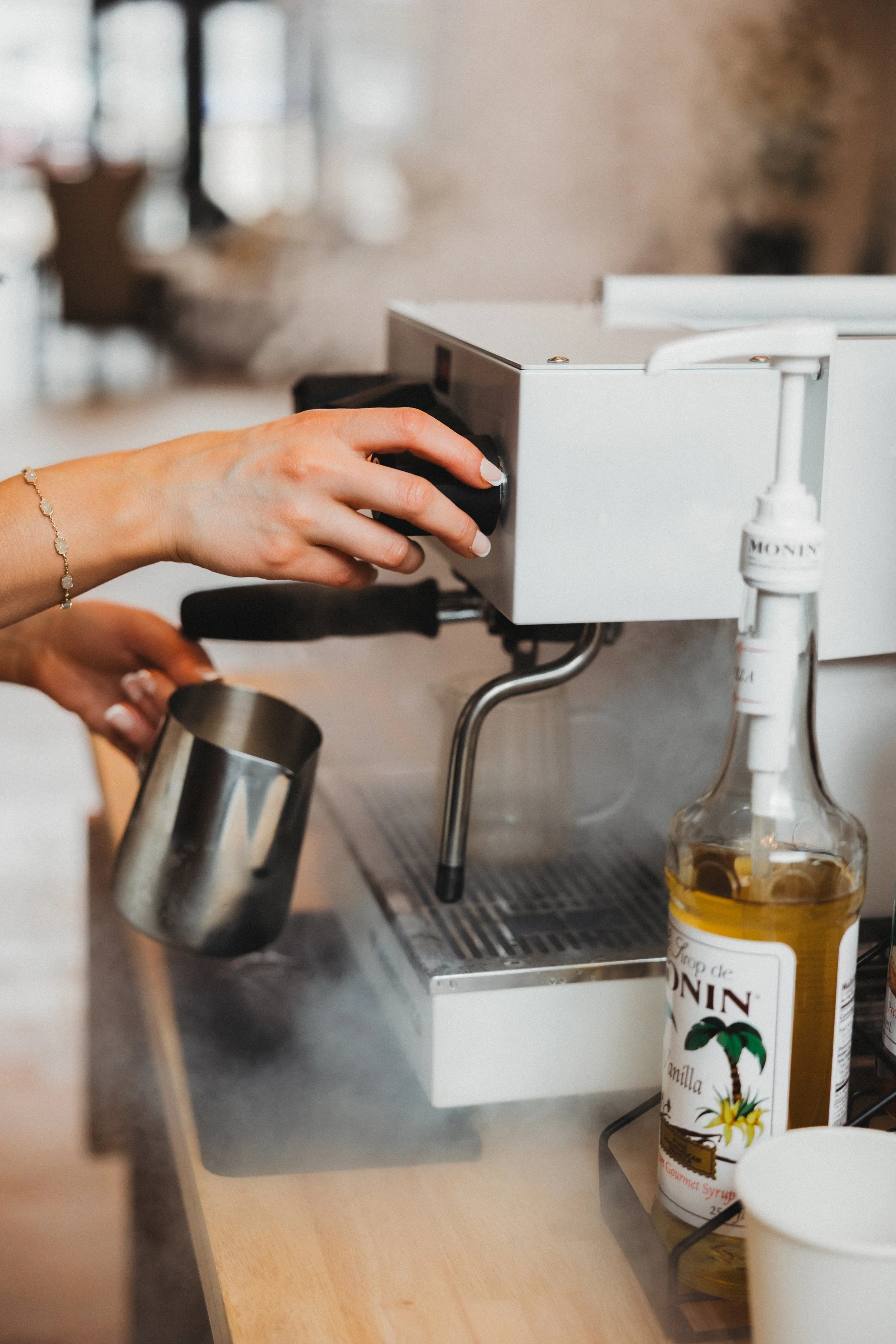 Person making coffee using a coffee machine, holding a small metal pitcher in one hand and adjusting the machine with the other, with a bottle of vanilla syrup nearby.