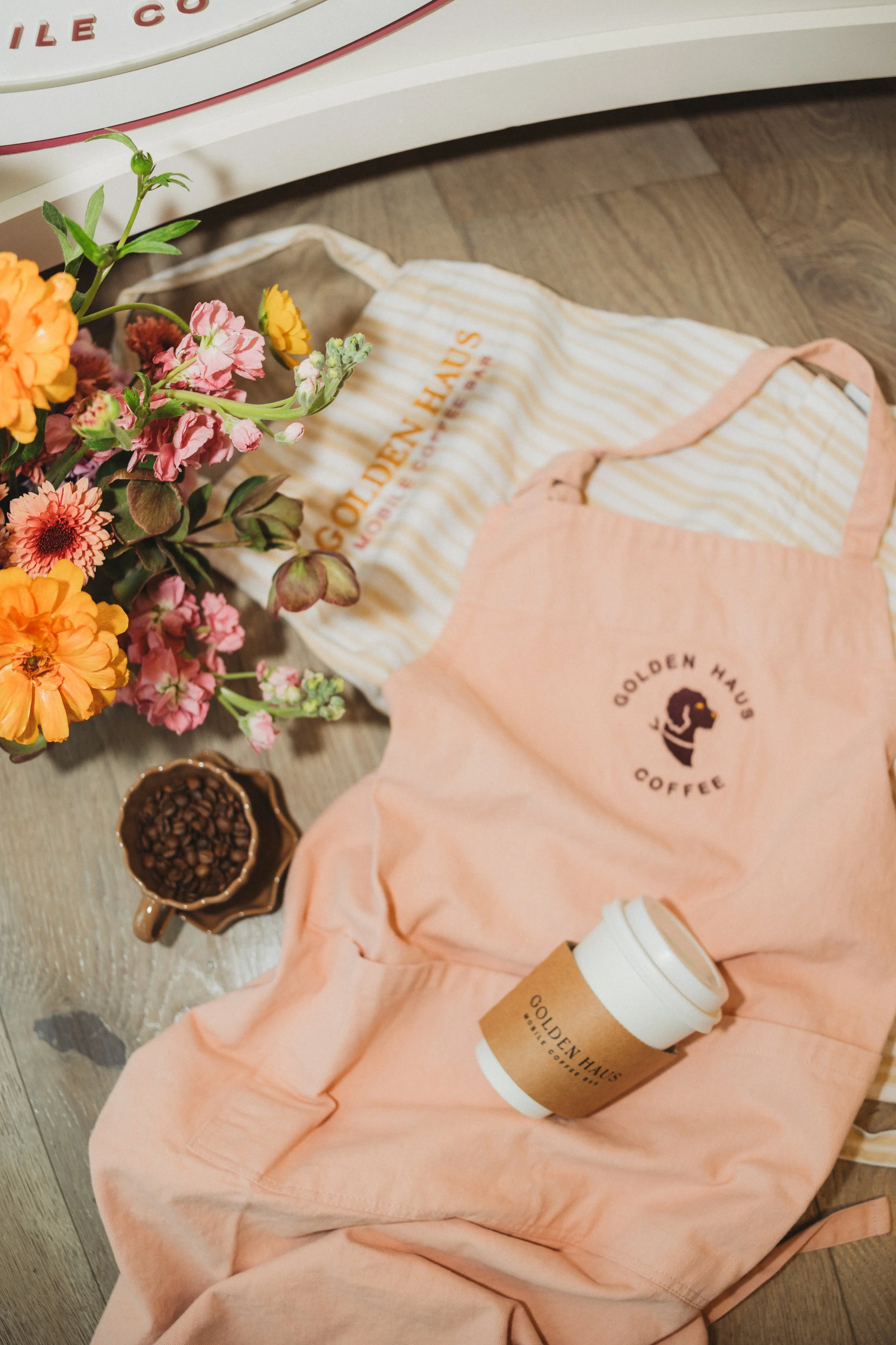 Flowers, coffee mug filled with coffee beans, paper coffee cup, and pink apron with 'Golden Haus Coffee' logo, arranged on a wooden floor.