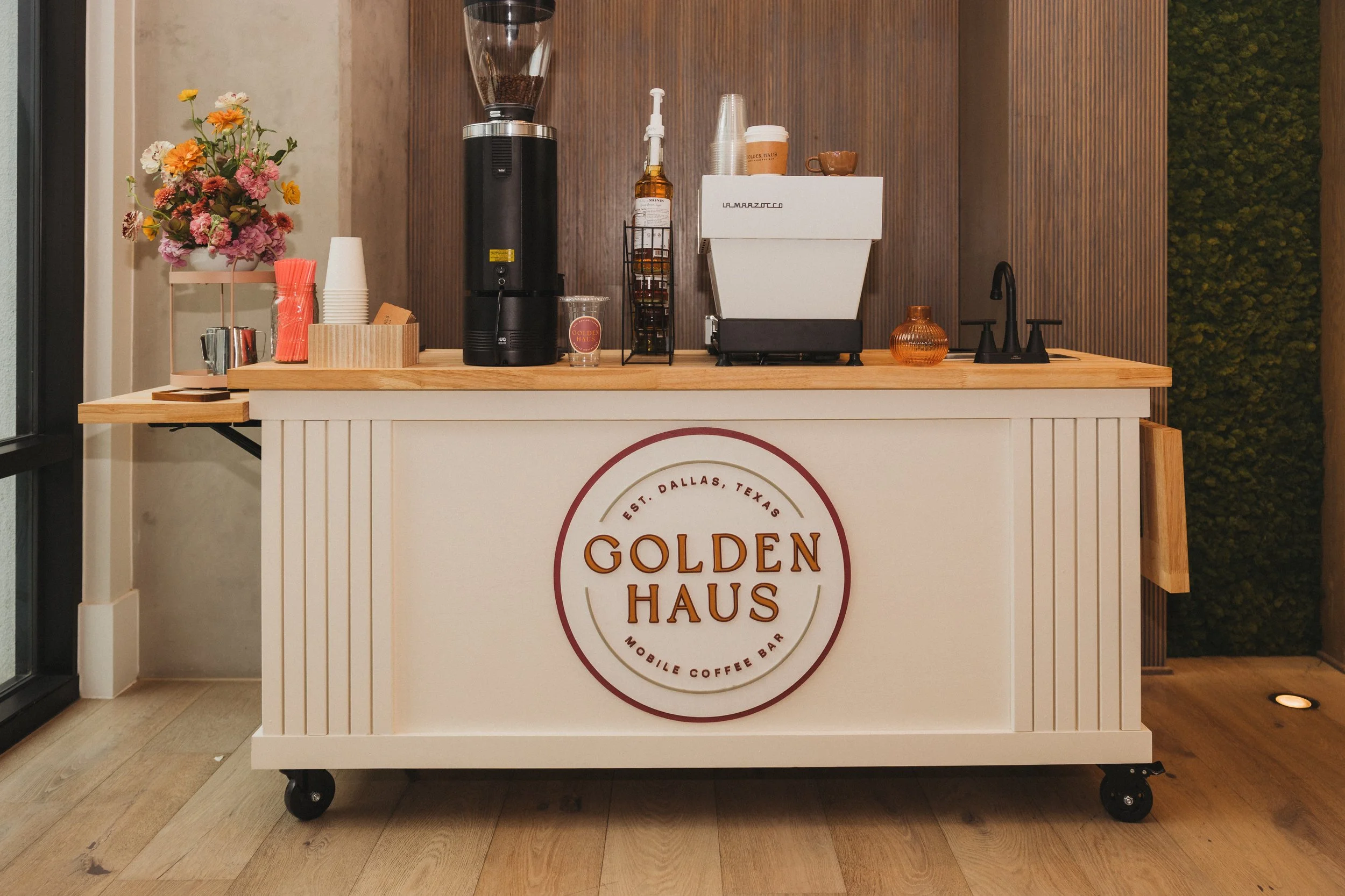 Mobile coffee bar with a white base, wooden top, and wheels. A large circular sign on the front reads "Golden Haus, Est. Dallas, Texas, Mobile Coffee Bar." The bar has a coffee grinder, a coffee machine, cups, a flower arrangement, and other items on top, with wood-paneled and green decorative backgrounds.