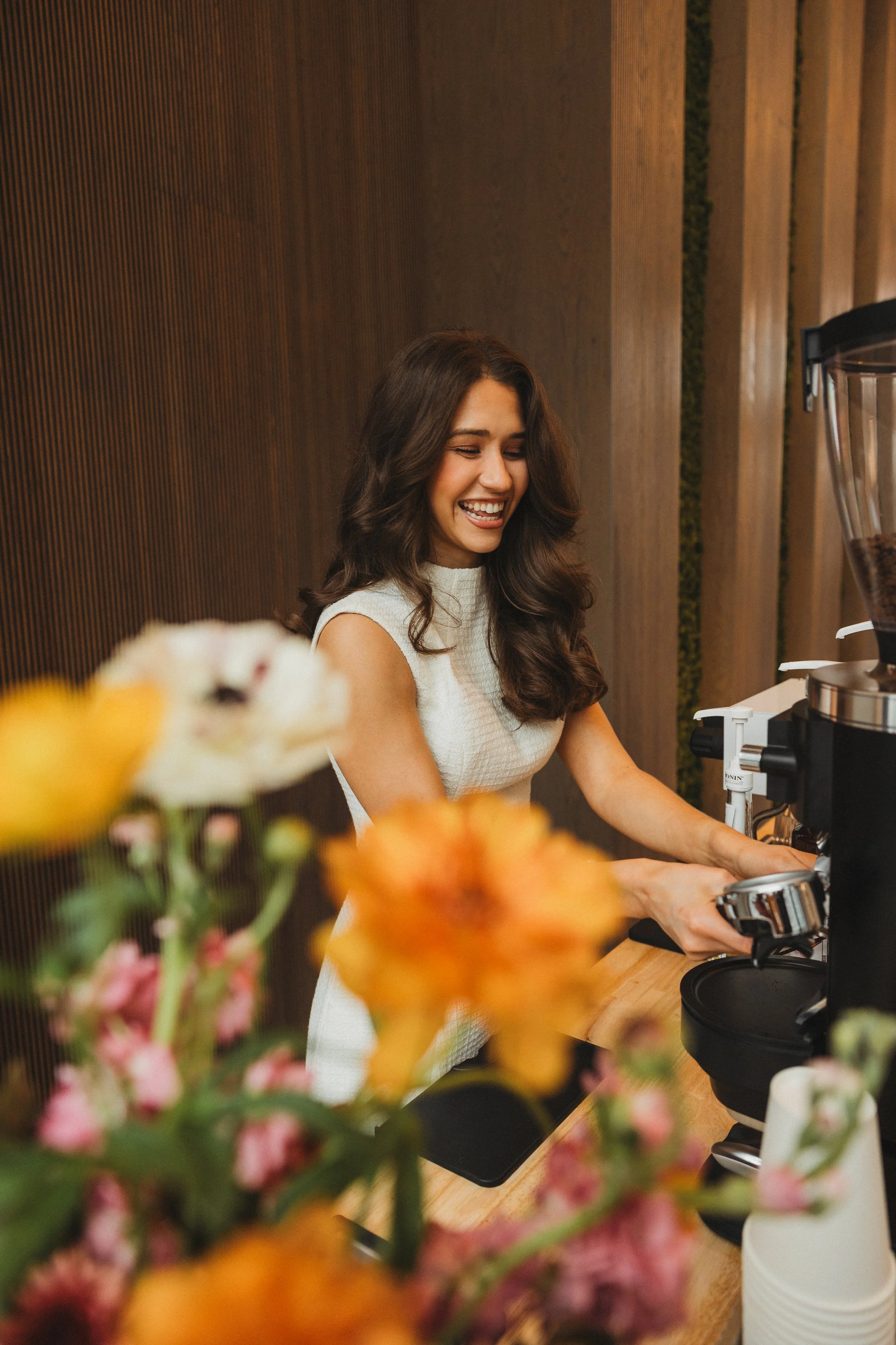 A woman with long curly brown hair, wearing a sleeveless white top, smiling while making coffee at a coffee shop counter with flowers in the foreground and a wooden wall in the background.