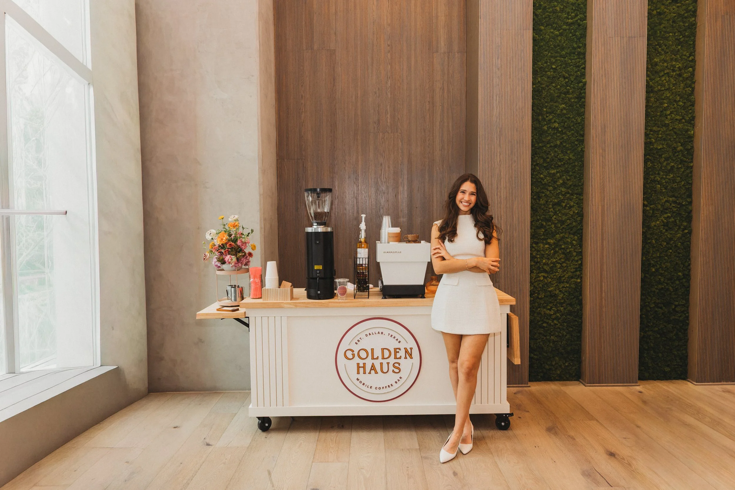 A smiling woman in a white dress standing in front of a coffee cart in a modern indoor setting, with a wooden and moss wall behind her and a large window to her left.