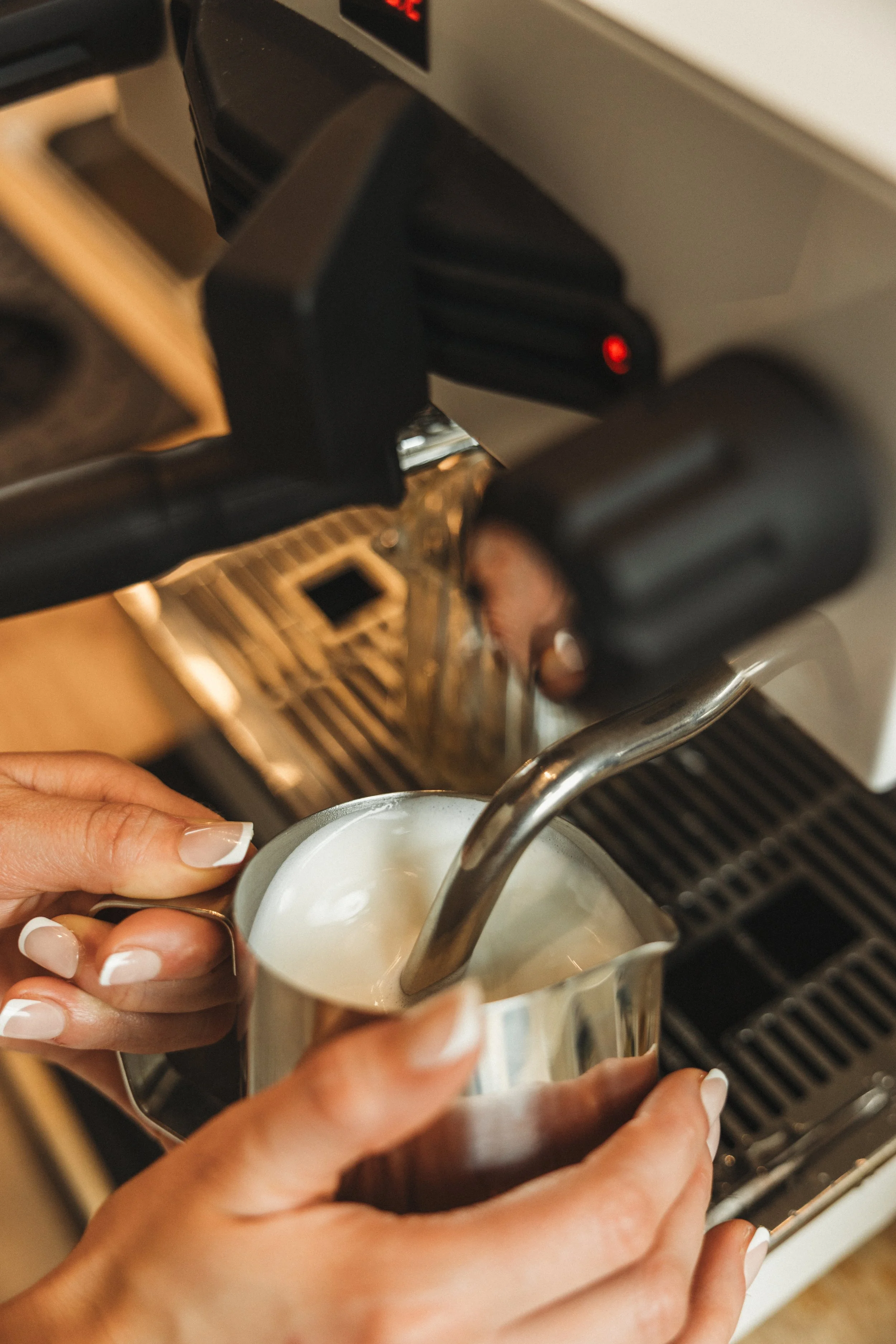 A person pours milk from a stainless steel pitcher into a coffee machine to make espresso.