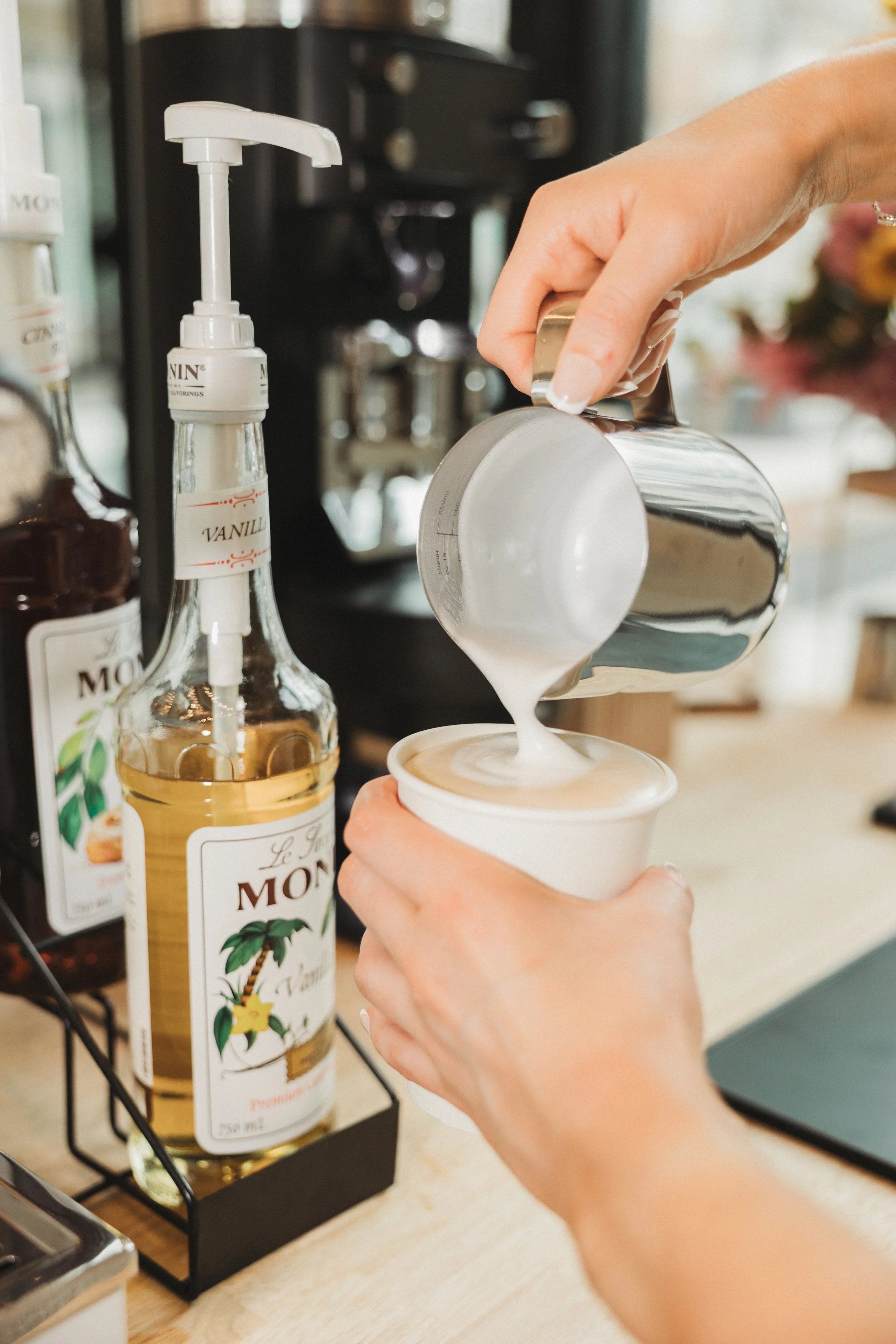 A person pouring milk into a coffee drink in a paper cup, with vanilla syrup in the background.
