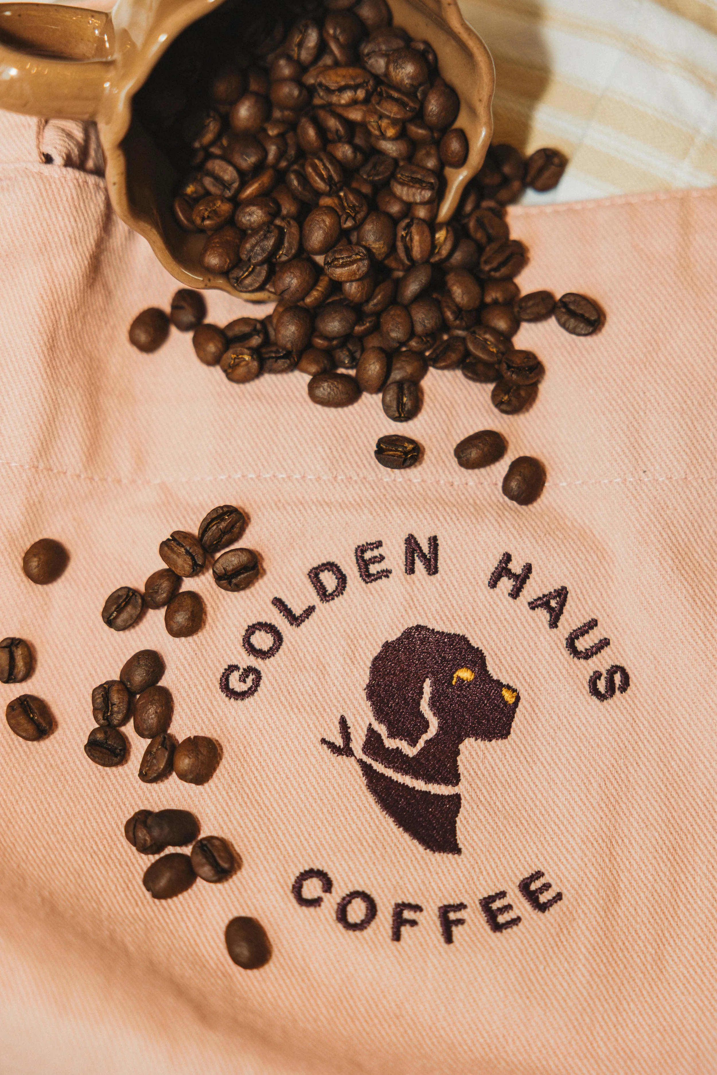 Coffee beans spilling out of a mug onto a pink cloth with the logo 'Golden Haus Coffee' and a dog silhouette.