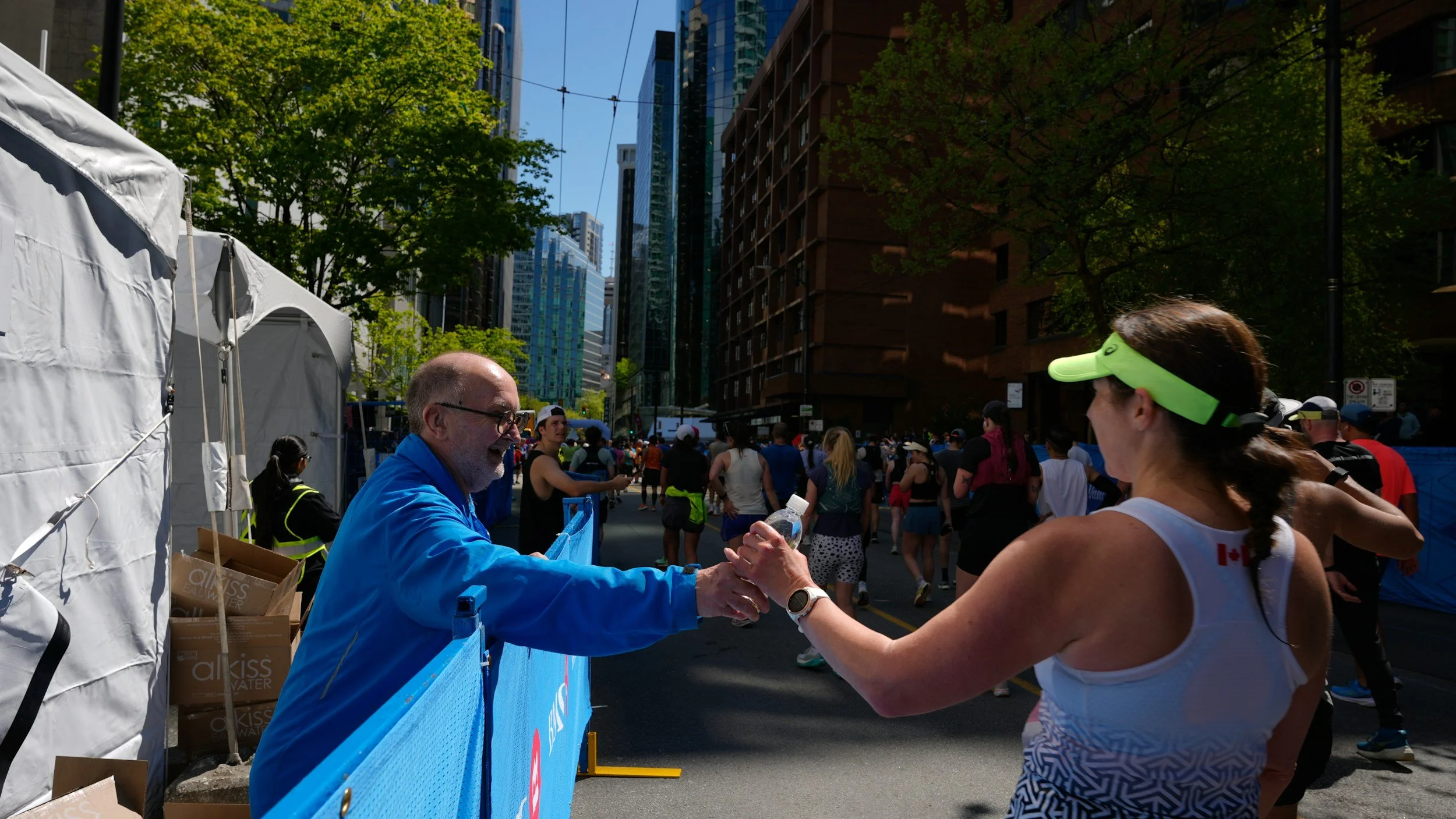 A man in a blue jacket and glasses is handing a water bottle to a woman in a white tank top with a neon green visor at an outdoor event in a city. The street is crowded with people, with tall buildings and trees in the background.