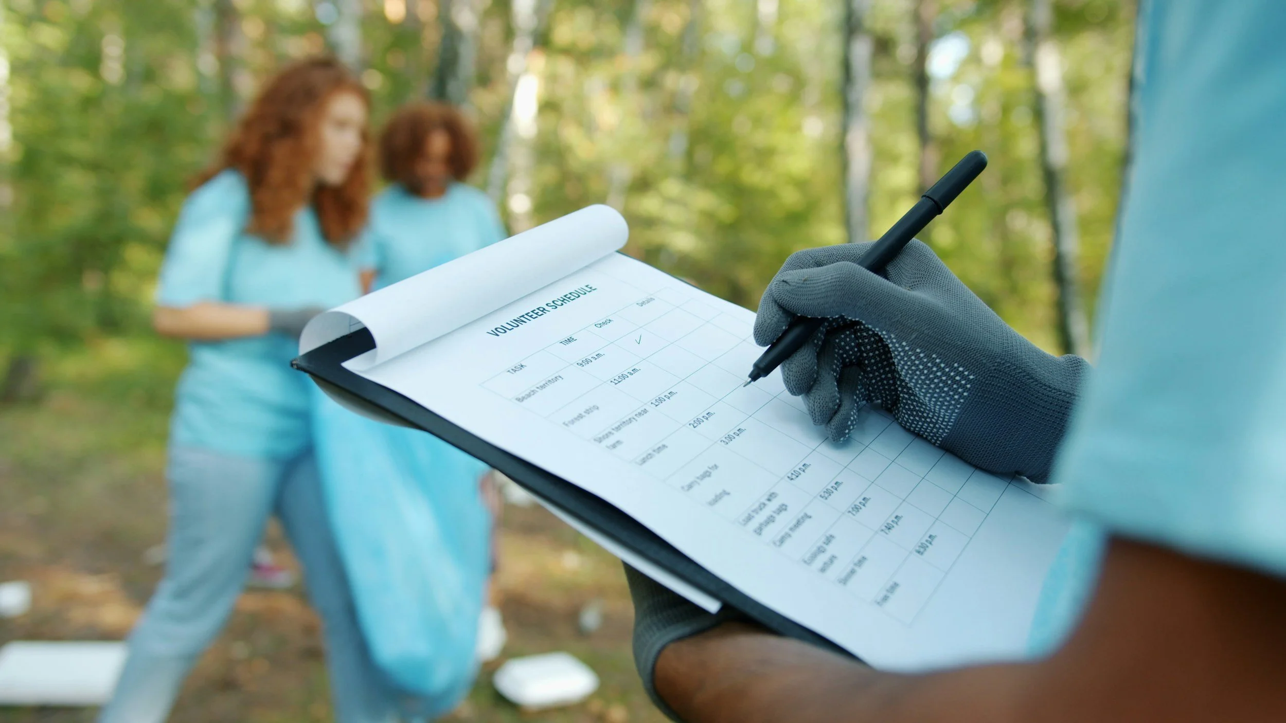 Person wearing gloves filling out a volunteer schedule on a clipboard while two women, dressed in blue, volunteer in a forested outdoor setting.