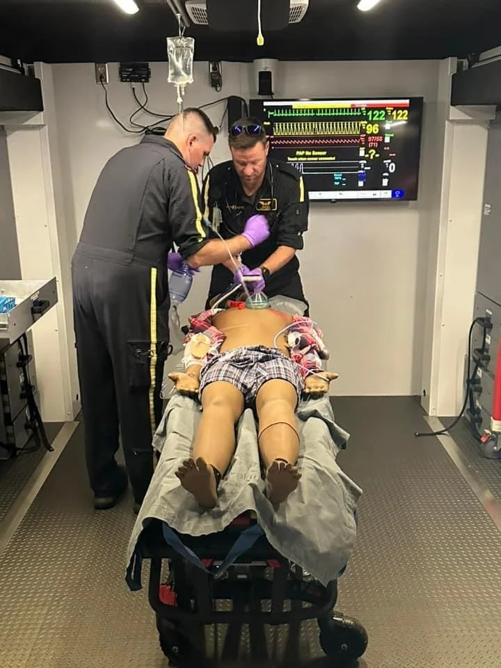 Two medical professionals attending to a patient on a stretcher in an ambulance or emergency room, with vital signs displayed on a monitor in the background.