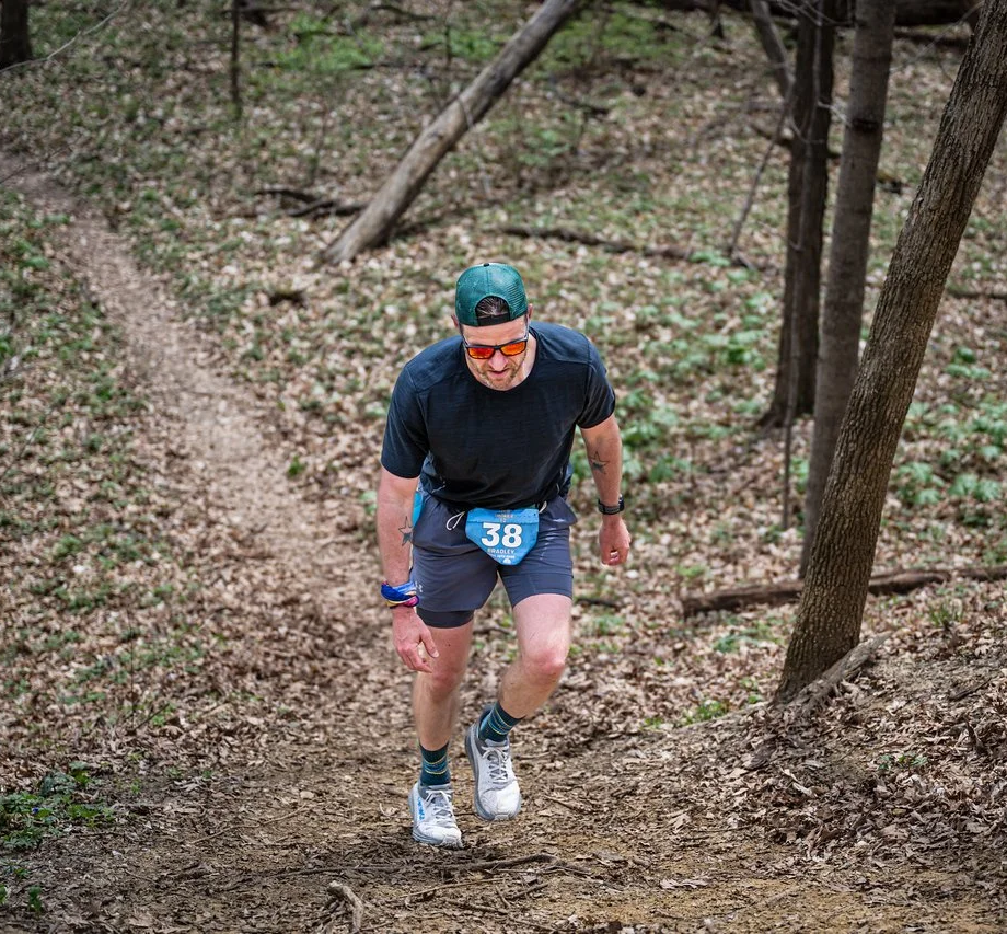 A man running on a dirt trail through a wooded area, wearing a black T-shirt, shorts, a green cap backwards, sunglasses, and a race bib numbered 38.