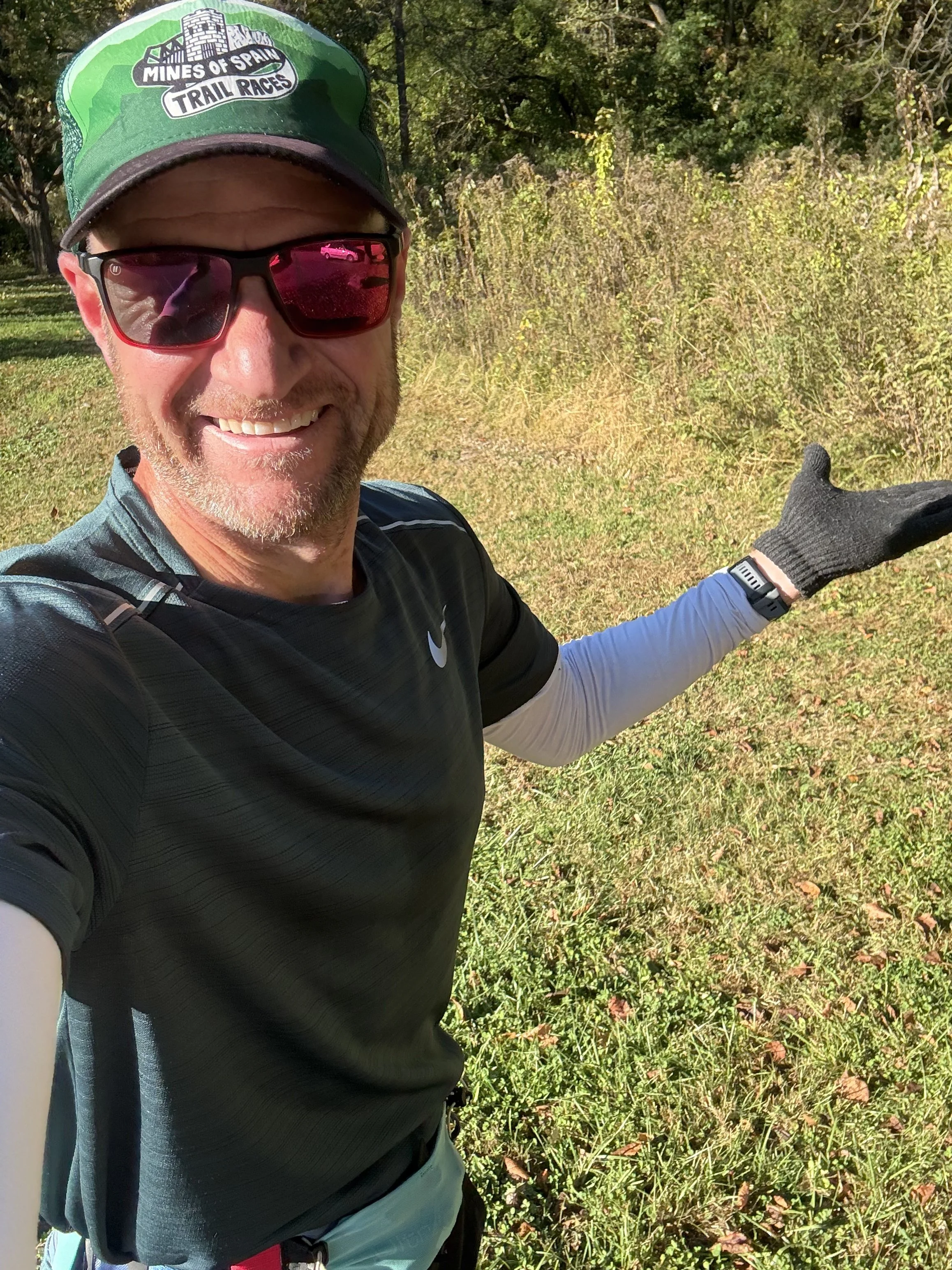 Man taking a selfie outdoors during a trail race, wearing sunglasses, a green cap, a black Nike shirt, a white long-sleeve shirt underneath, a black running glove, and a fitness tracker, with trees and a grassy area in the background.