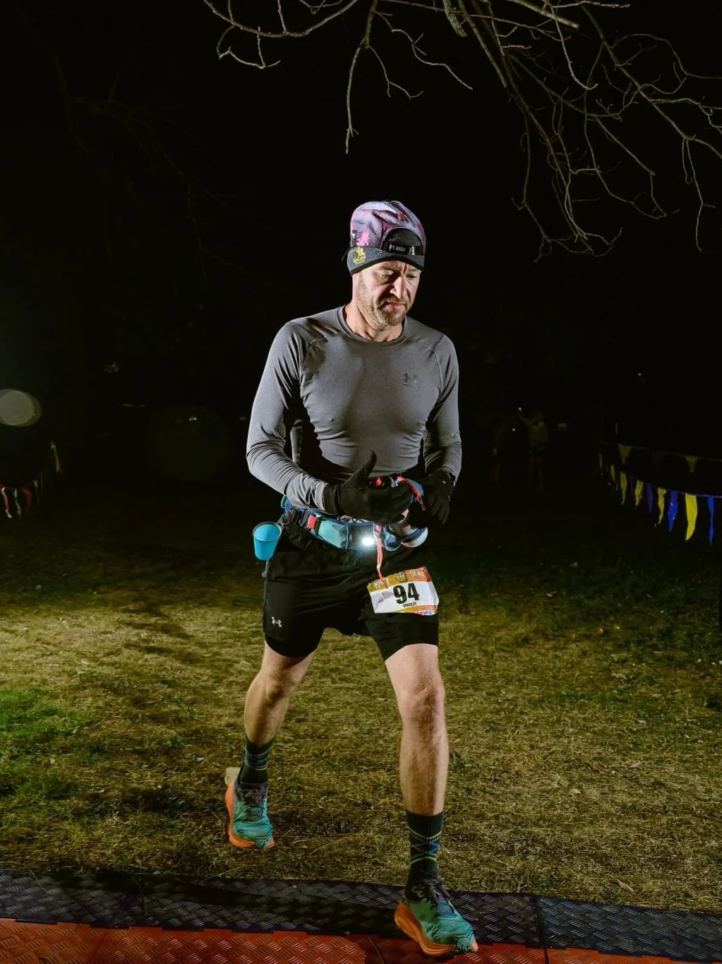 A male runner in athletic gear, wearing a hat, gloves, and a bib number 94, is running at night on a trail illuminated by a light source, with trees and yellow and purple flags in the background.