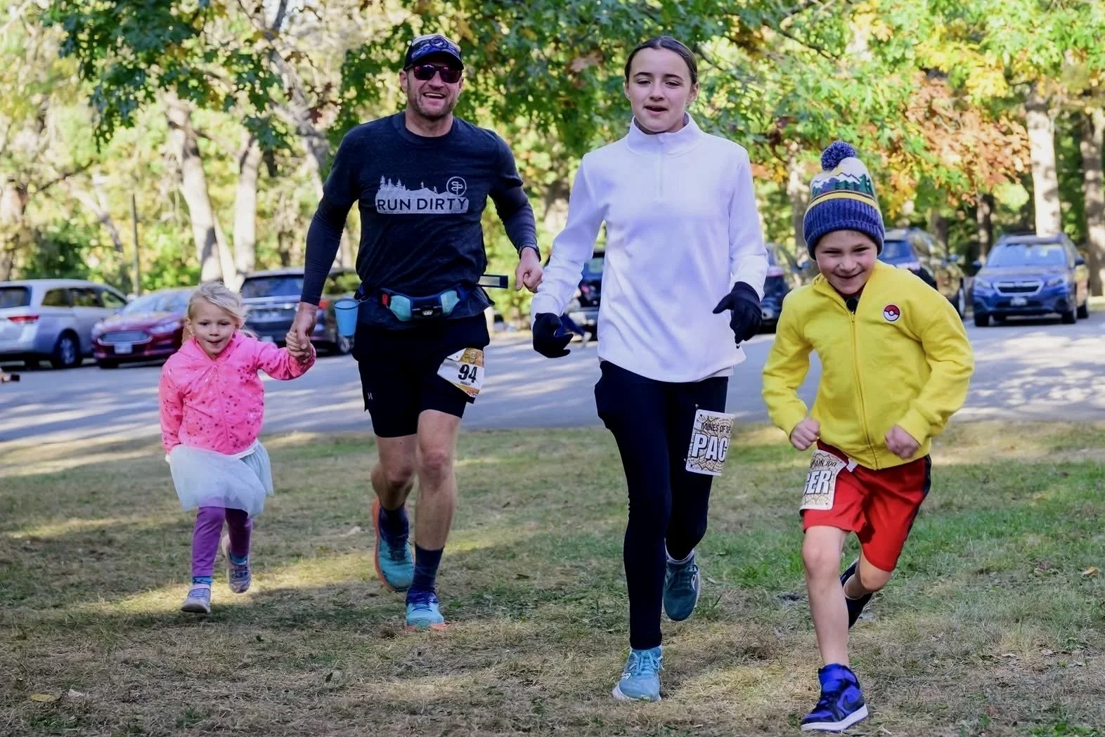 A group of people participating in a running event in a park during autumn, including two children, a woman, and a man. The children are smiling and dressed warmly, with one in a yellow jacket and blue hat, and the other in pink and purple. The man and woman are wearing running gear with race numbers, surrounded by autumn trees and parked cars.