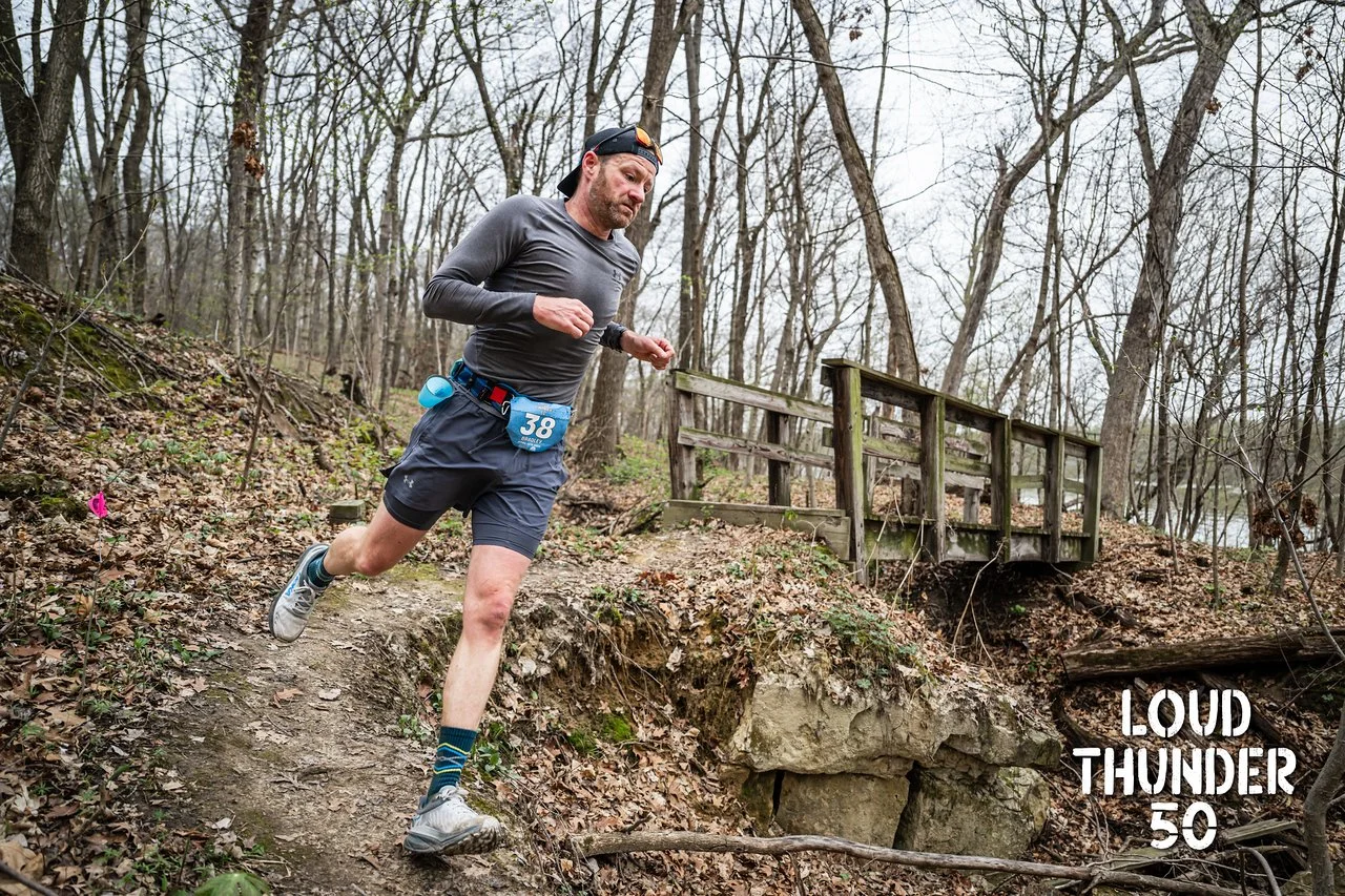 A man running downhill on a trail in a wooded area during a race, wearing a gray shirt, black shorts, and a black cap backward, with a bib number 38 and a water bottle attached to his shorts, with the words "LOUD THUNDER 50" overlayed on the lower right corner of the image.
