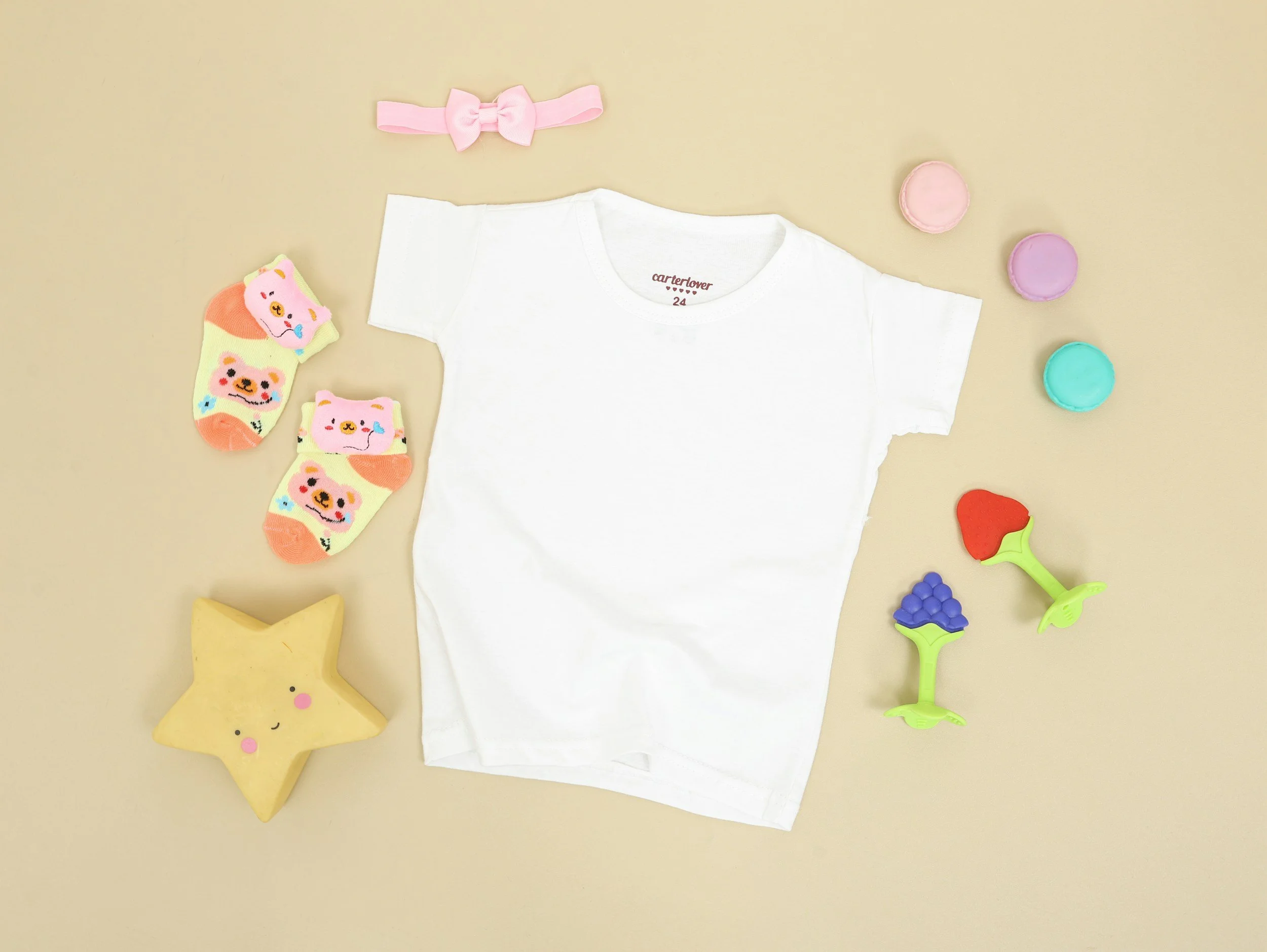 Flat lay of a white children's T-shirt, pink bow headband, colorful socks with bear and cat faces, star-shaped toy with a smiling face, and various small plastic toys on a beige background.