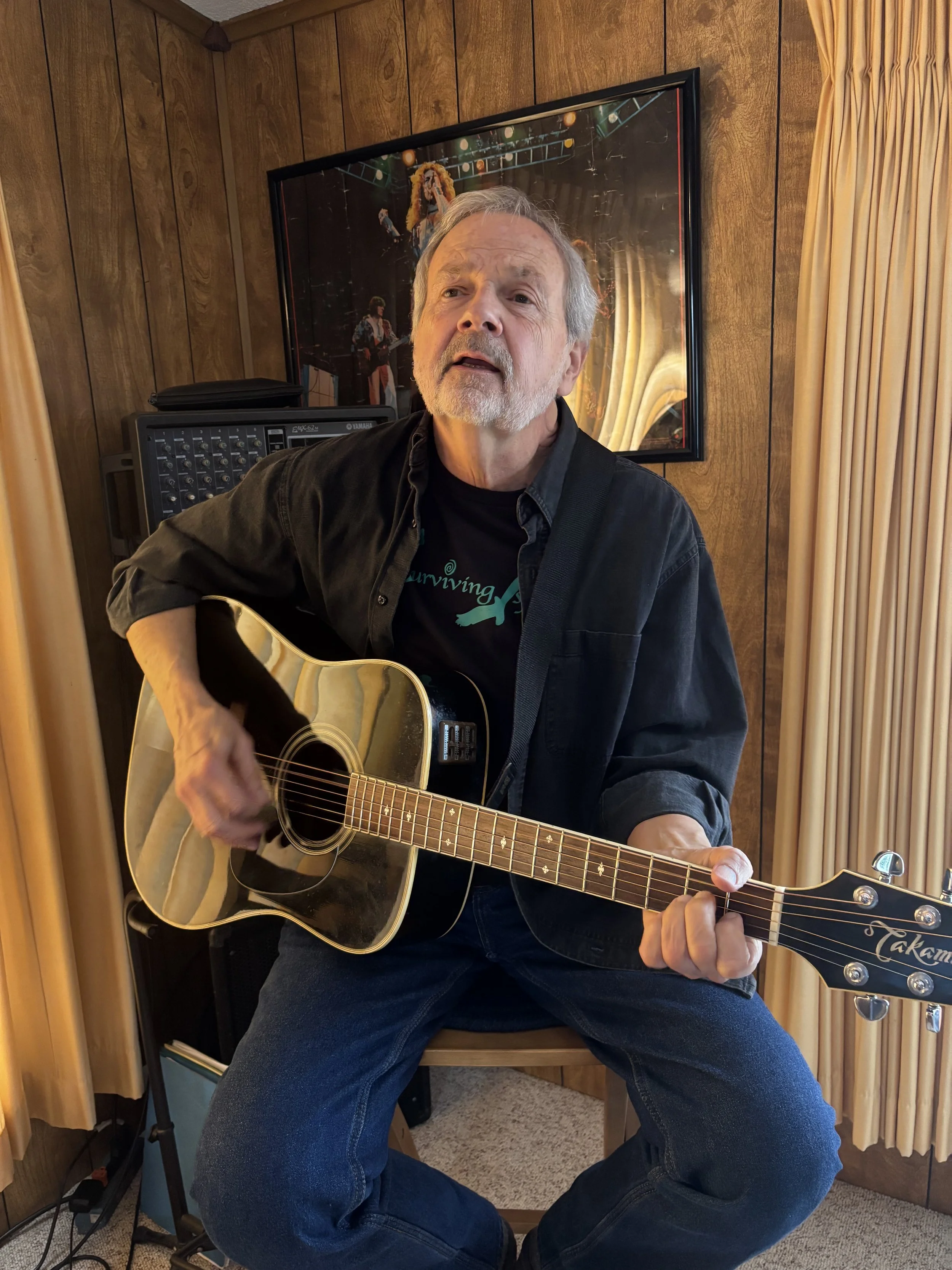 An older man with gray hair and a gray beard playing an acoustic guitar in a room with wood-paneled walls, a framed picture of a band on the wall behind him, and beige curtains on the window.