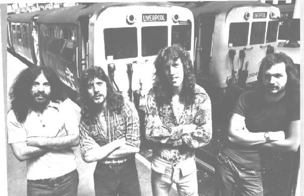 Four people with long hair and beards standing in front of train cars labeled 'Liverpool'