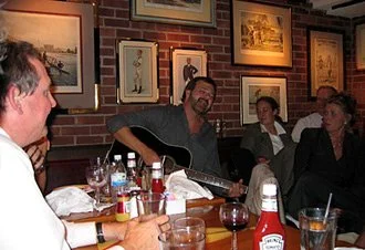 Group of people sitting around a table in a restaurant, with one person playing guitar, and others listening and talking, framed pictures on brick wall in background.