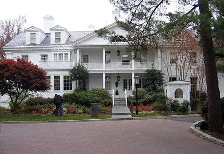 Large white mansion with multiple porches, surrounded by trees and shrubs, and a brick walkway leading to the entrance.