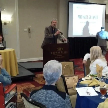 A man is giving a presentation at a conference, standing behind a podium with a screen displaying his name, Michael Skinner. Several audience members are seated at tables, with some looking toward him.
