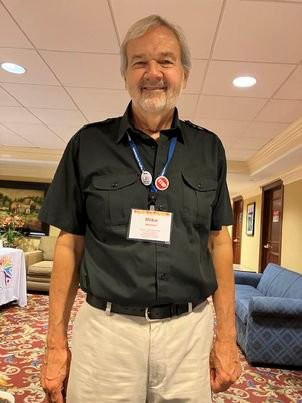 An older man with gray hair and a beard smiling, wearing a black short-sleeved shirt, beige pants, and a conference badge around his neck, standing in a hotel conference room.