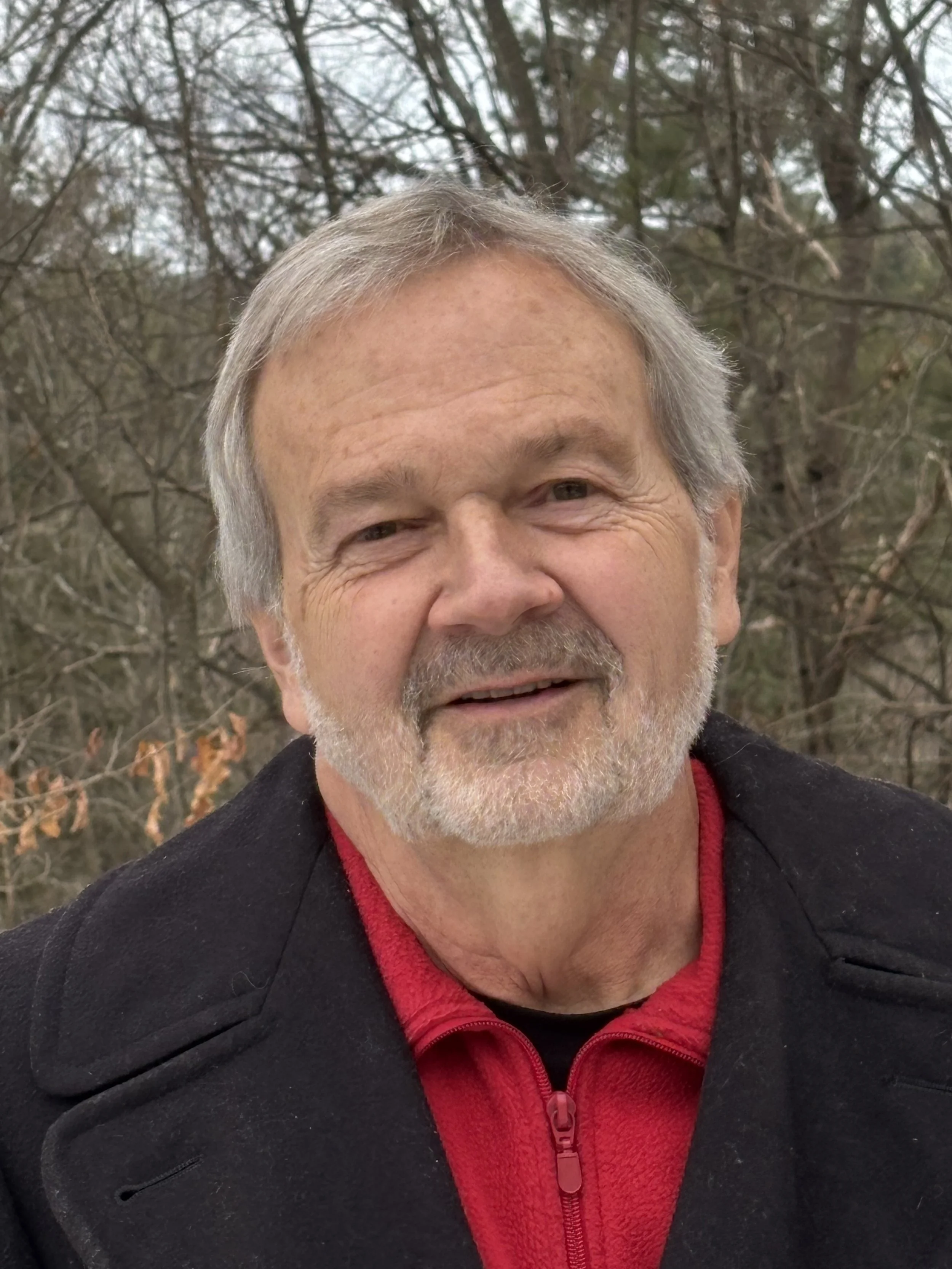 A smiling middle-aged man with gray hair and a beard, wearing a black jacket over a red shirt, outdoors with leafless trees in the background.