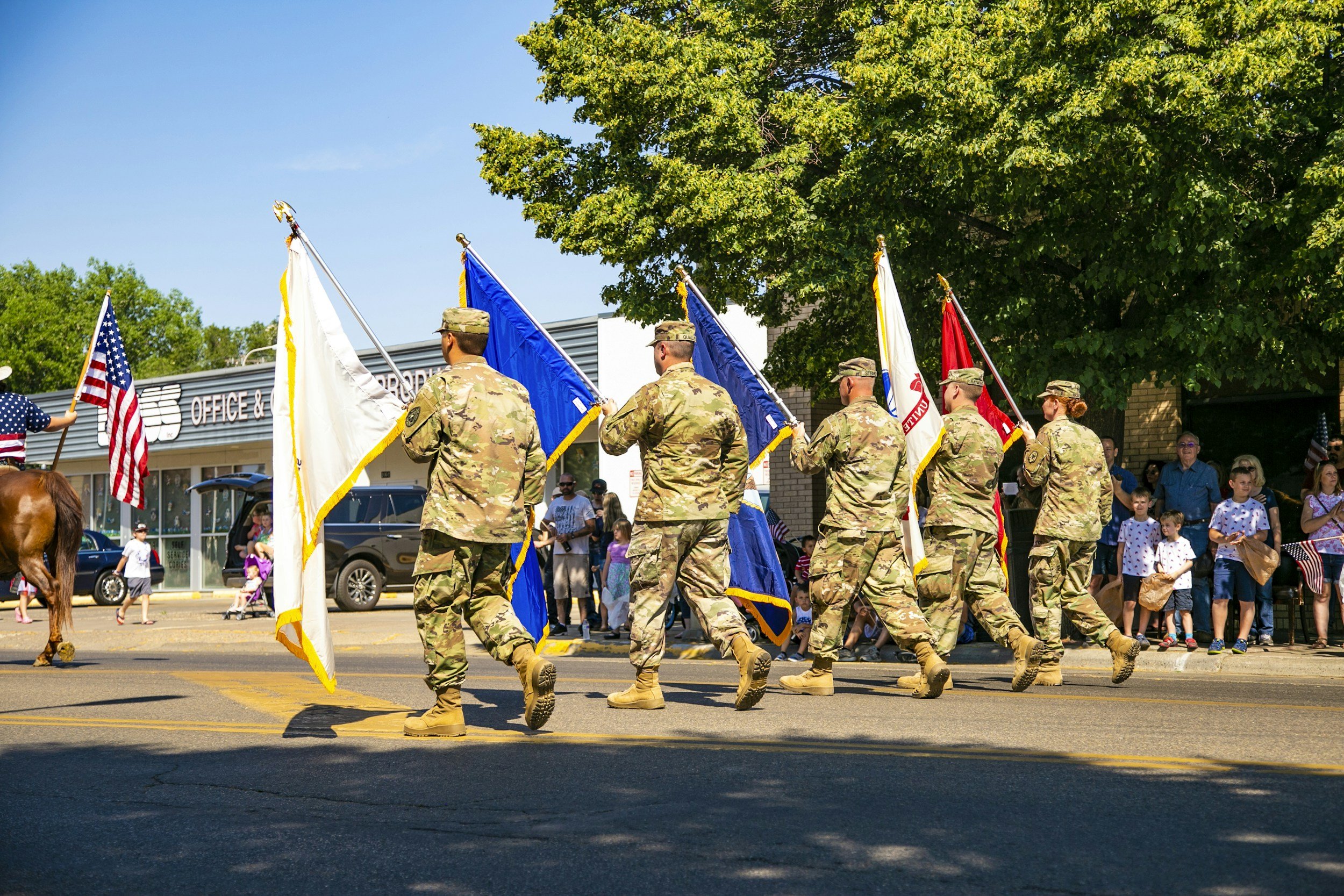 Soldiers marching in a parade carrying flags, with spectators watching from the sidewalk and a building in the background.