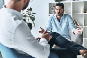 A man in a white shirt and a man in a light blue shirt are sitting and talking in an office or therapy setting, with one gesturing with hands.