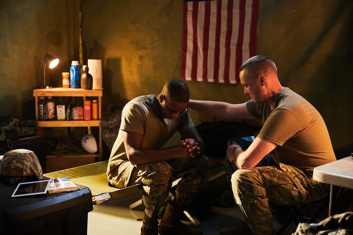 Two soldiers in military uniforms sitting with heads bowed and hands clasped, engaged in prayer inside a dimly lit room with a US flag on the wall.