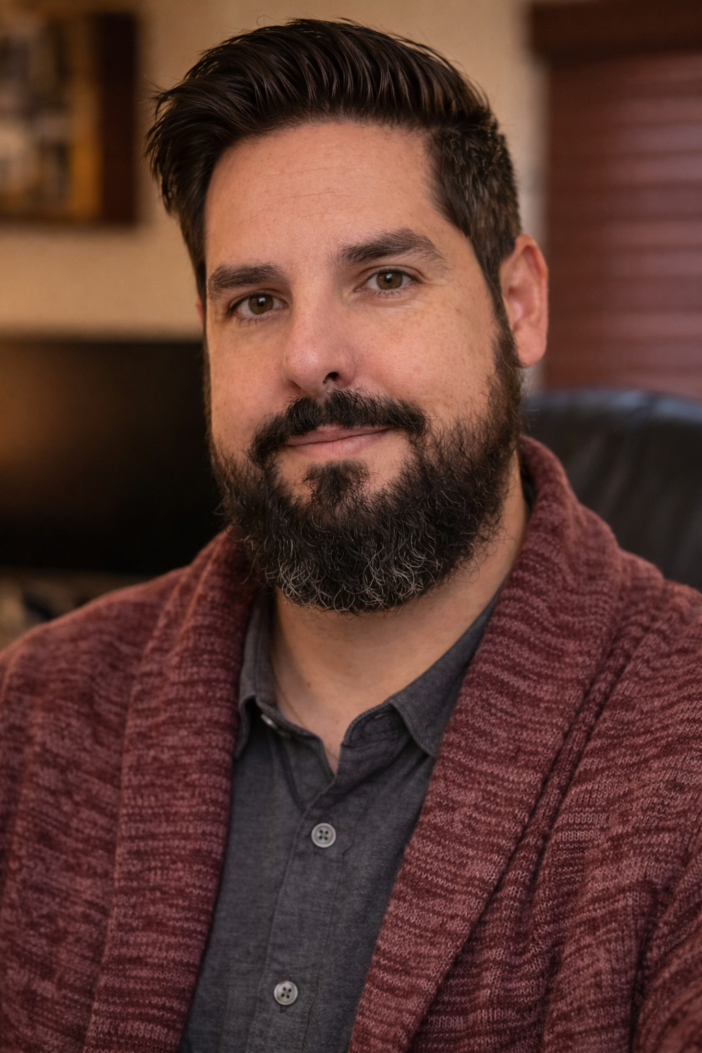 A man with a beard and moustache, wearing a dark shirt and a maroon sweater, smiling softly at the camera. The background features a cozy indoor setting with warm lighting.