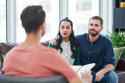 A woman and man sitting on a couch during a counseling session, facing a therapist with their backs to the camera.