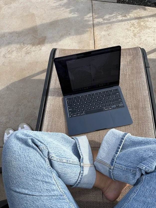 A laptop on a lounge chair, with a person sitting cross-legged wearing light blue jeans, taken from above.