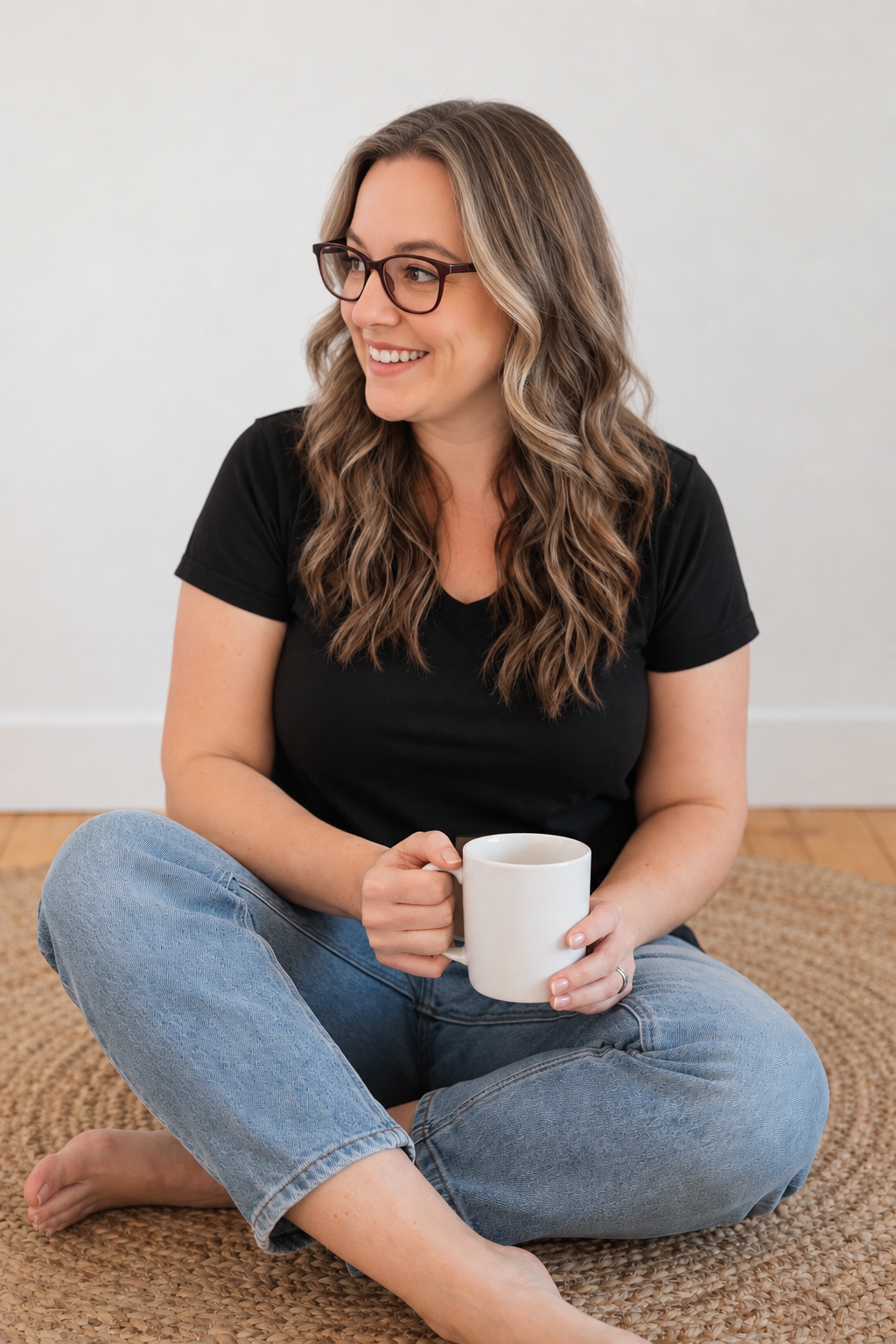 Rebecca, founder of Modern Shift Studio, with wavy hair and glasses, sitting and holding a white ceramic mug and smiling, wearing a black t-shirt and blue jeans.