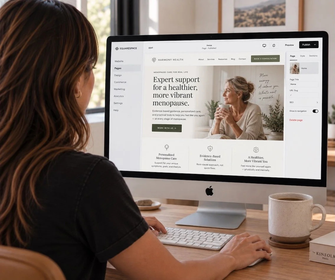 A woman using a computer to design a website for a health care service focused on menopause support, with a mug of coffee and a book on a wooden desk.