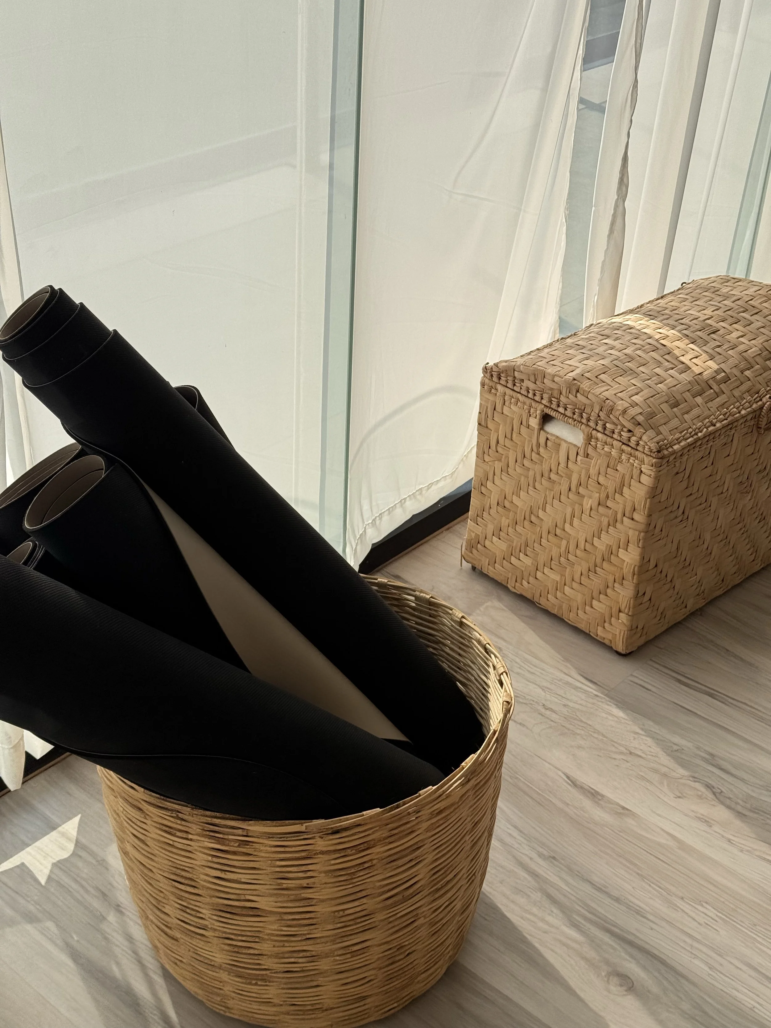 A wicker basket with a yoga mat inside, placed on a wooden floor near a glass wall with curtains.