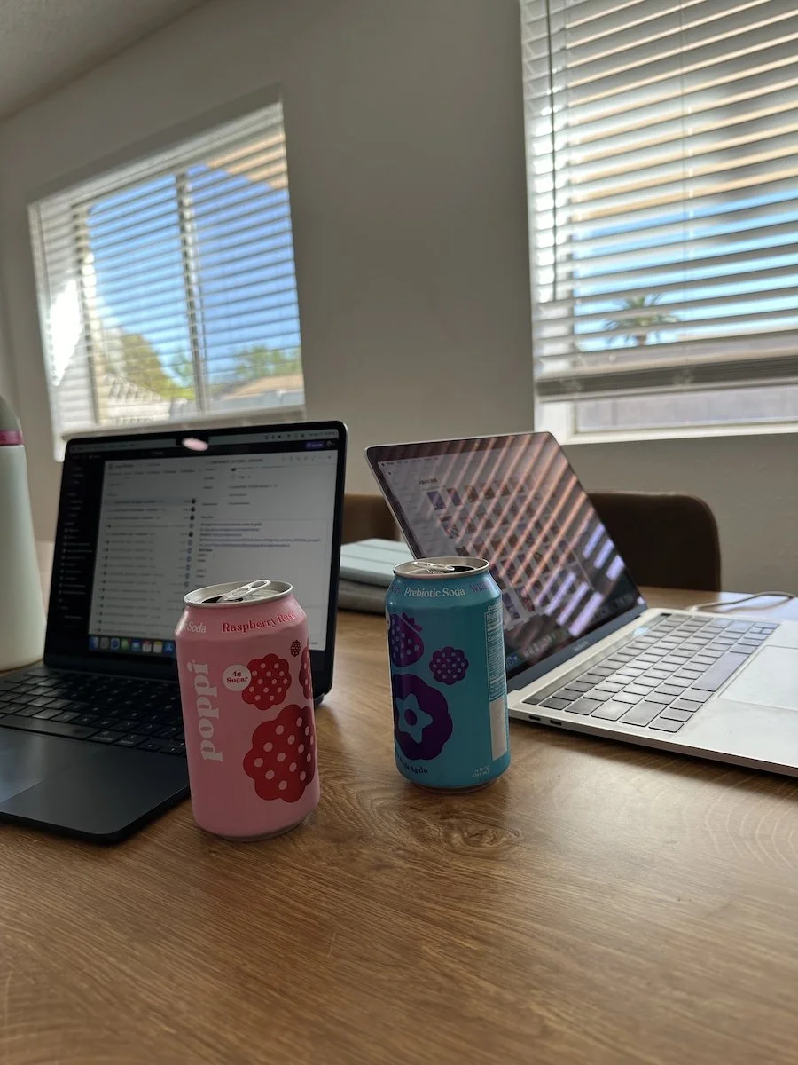A wooden kitchen table with two open laptops and two cans of poppi in front of a window with sunlight streaming in.