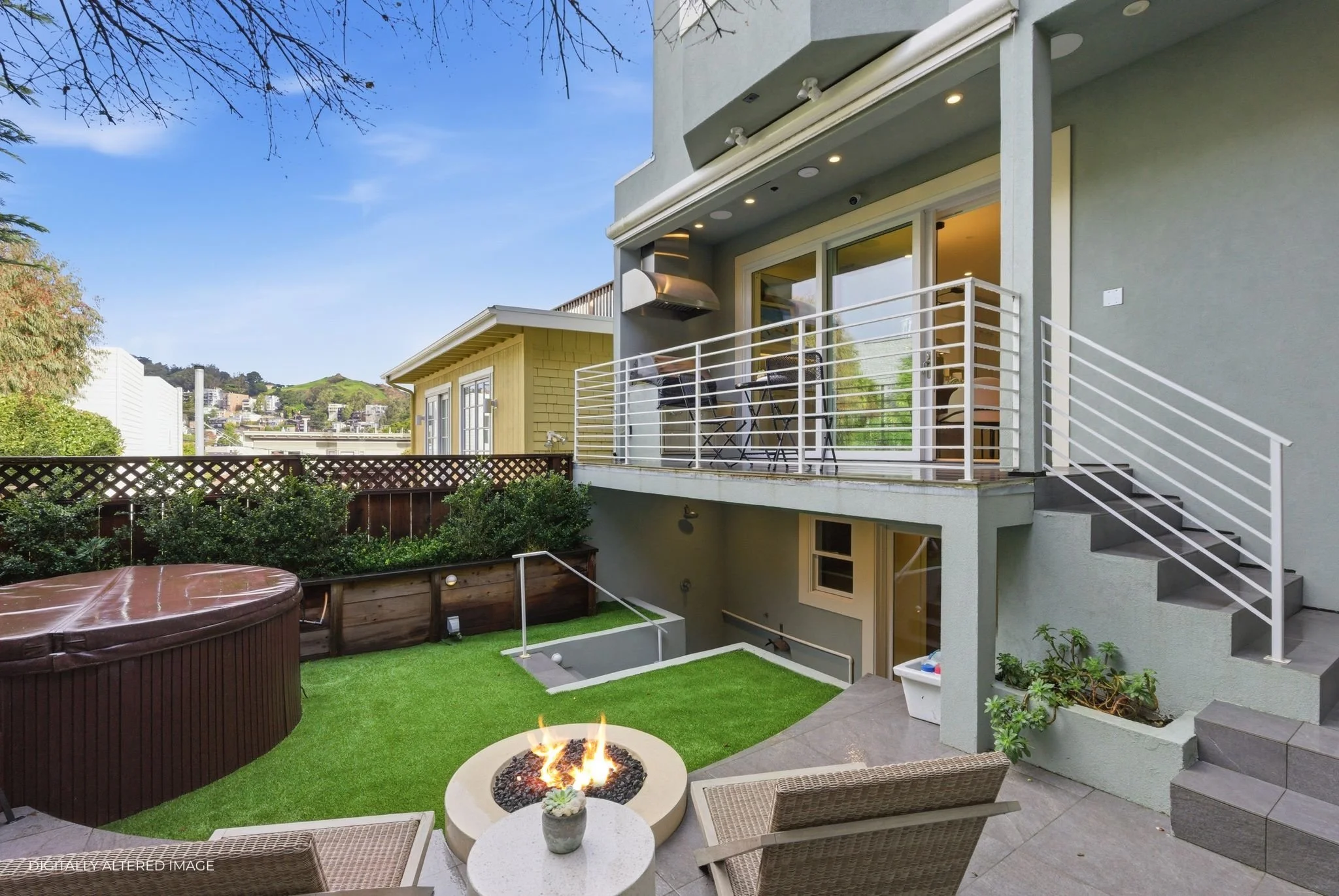 Backyard patio with fire pit, hot tub, outdoor seating, balcony with grill, and stairs, in a residential area with hills in the background.