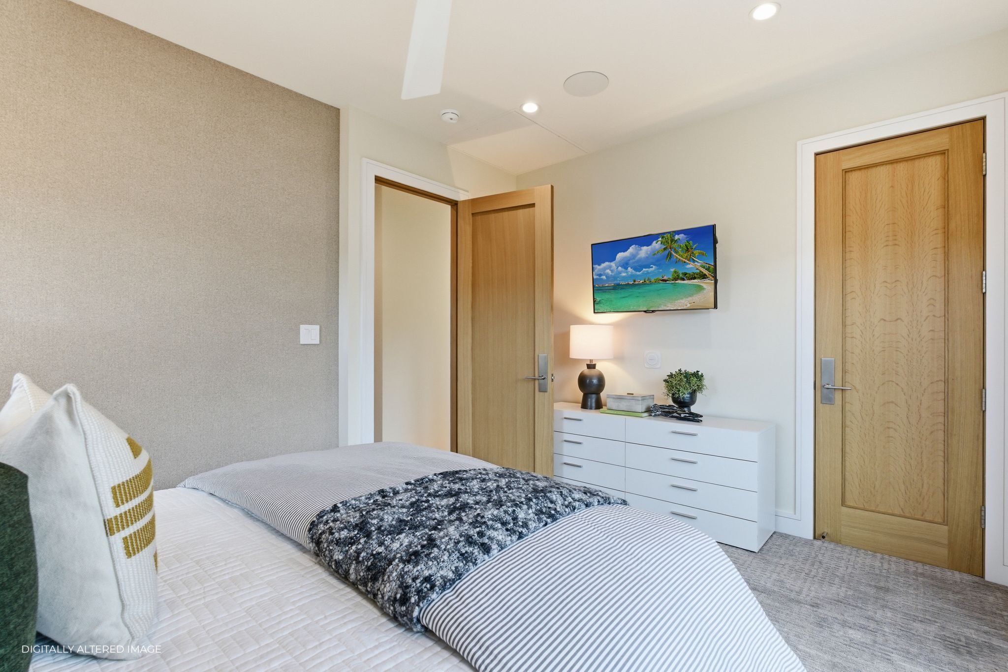 Modern bedroom with a bed, beige accent wall, wooden door, wall-mounted TV displaying a beach scene, white dresser with a lamp, plant, and books, with neutral-colored carpet.