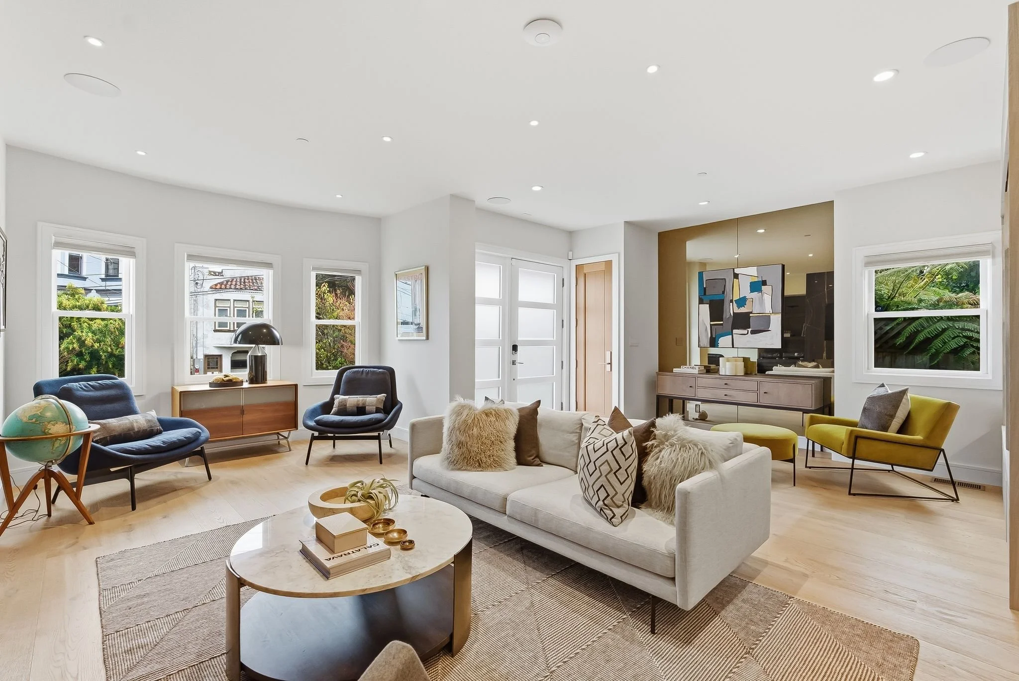 Bright living room with a white sofa, black and green chairs, a wooden sideboard, and large windows with greenery outside.