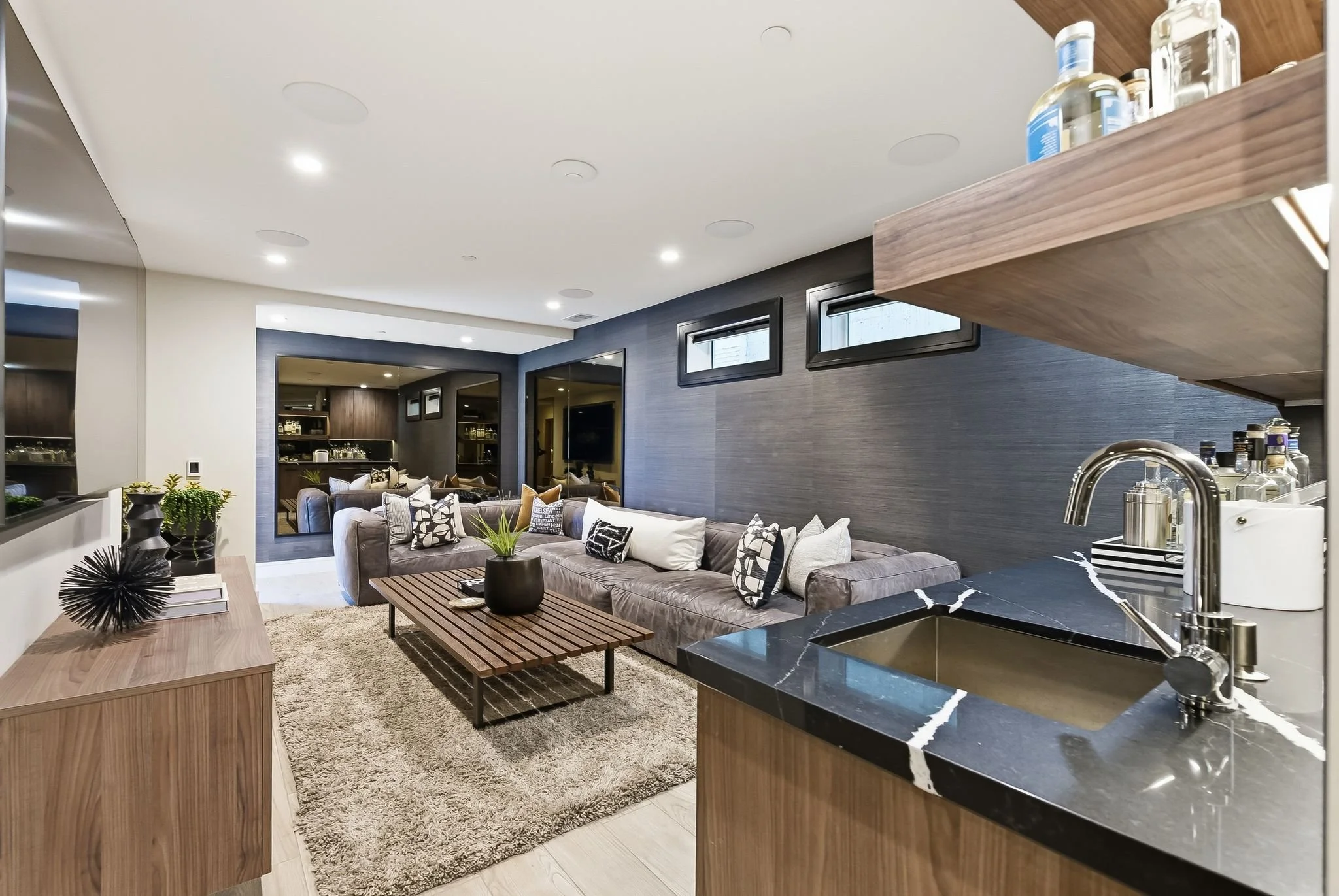 Modern living room with a sectional sofa, wooden coffee table, and black marble kitchen counter, featuring dark accent wall and small high windows.