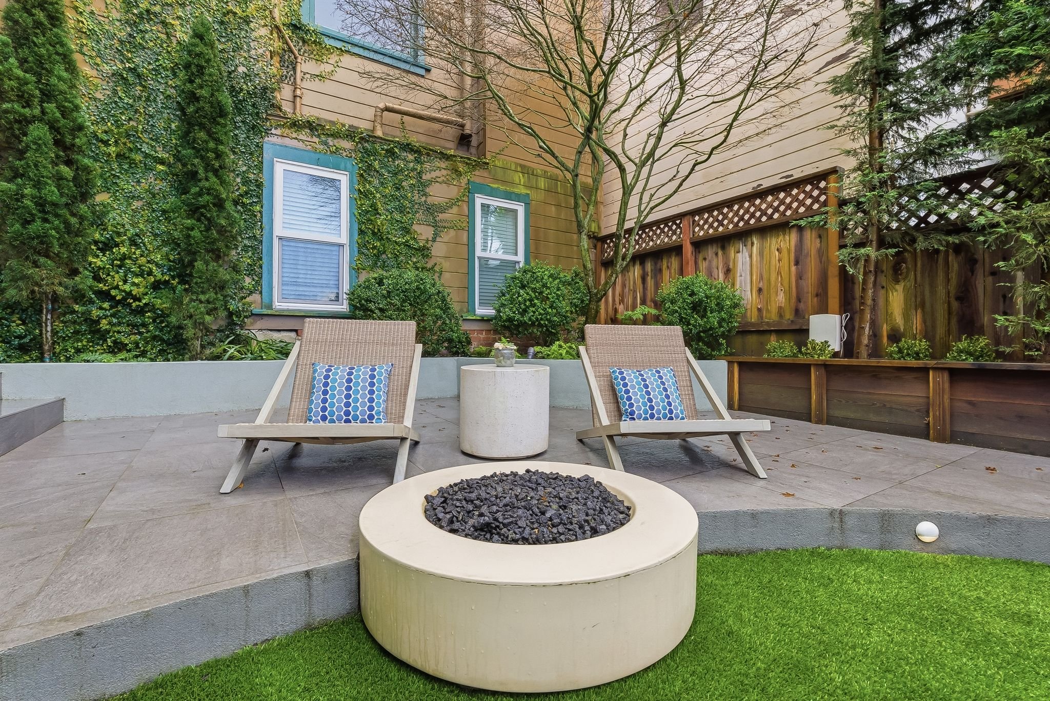 Outdoor backyard patio with two beige lounge chairs, a small white table, a fire pit filled with black stones, and a lush green lawn. The background includes a wooden fence, greenery, and a house with blue window trim.