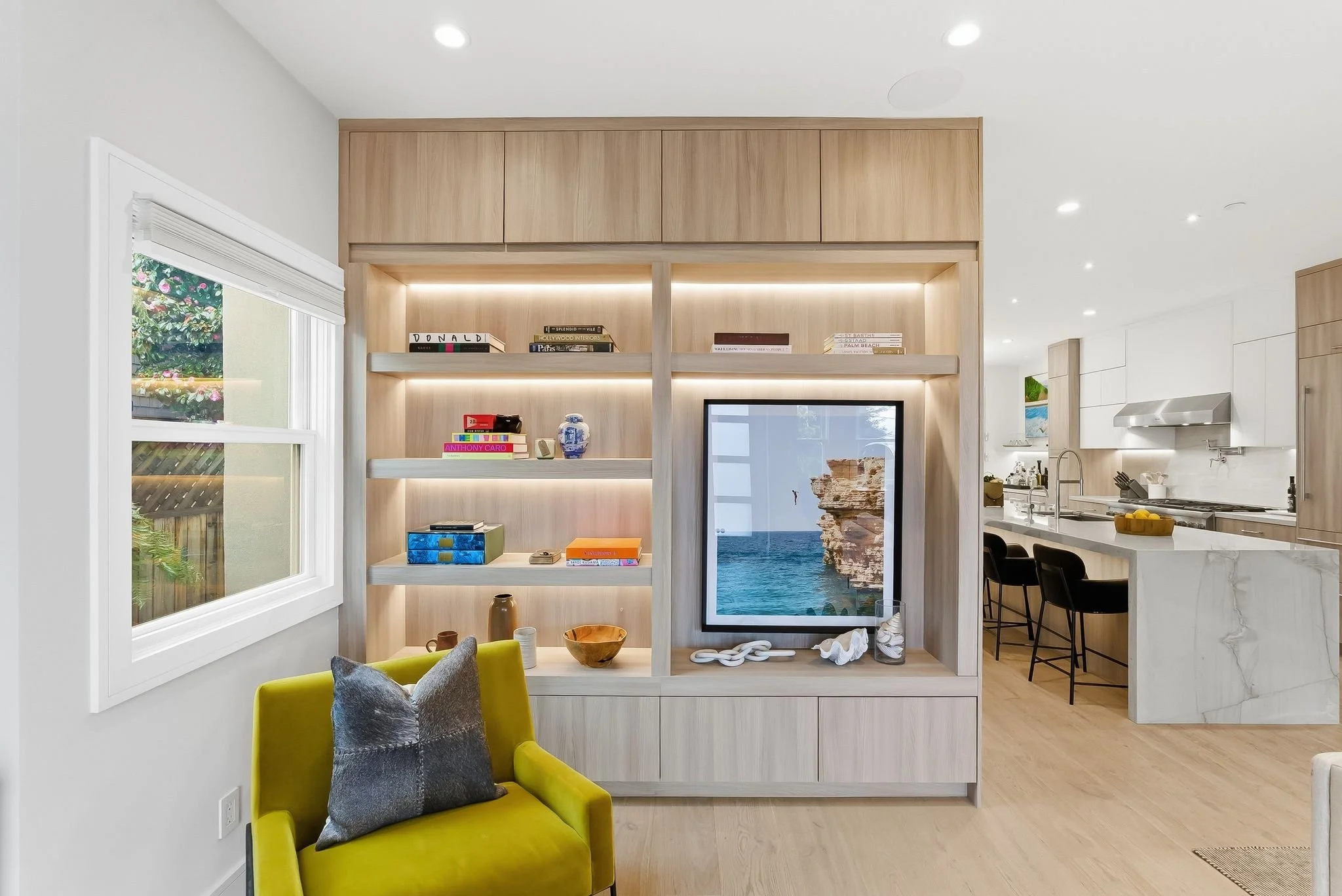 Modern living room with built-in wooden shelving unit, decorative objects, and a large framed sea cliff photograph. Kitchen in background with island, black bar stools, and stainless steel appliances.