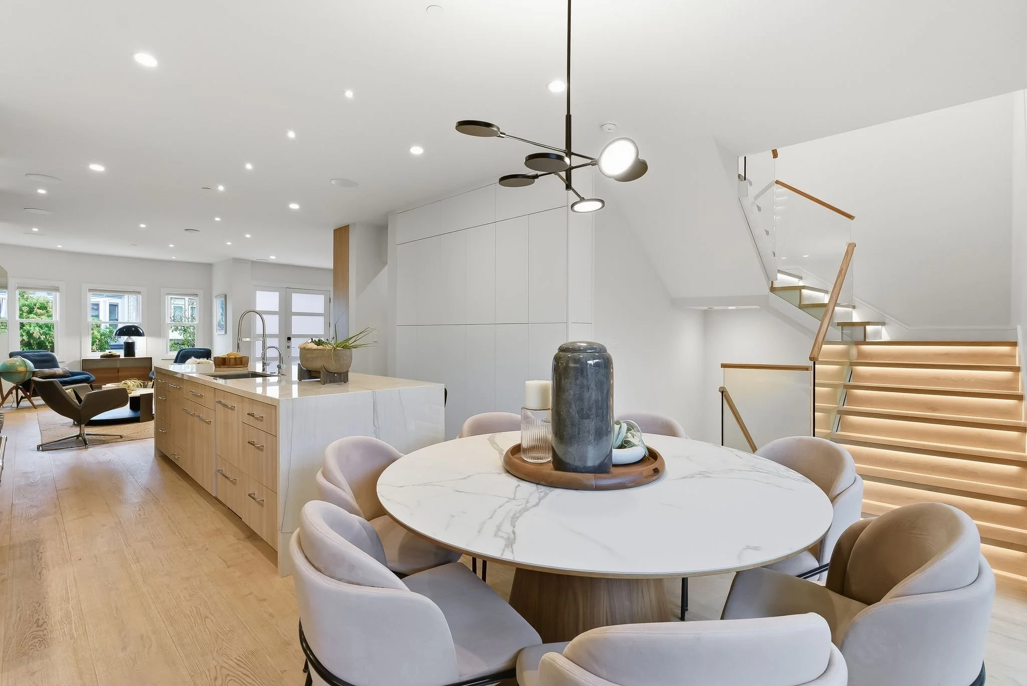 Modern open-concept living and dining area with white walls, light wood flooring, and minimalist furniture. Includes a round marble dining table with beige upholstered chairs, a kitchen island with a sink, and a staircase with wooden steps and lighti